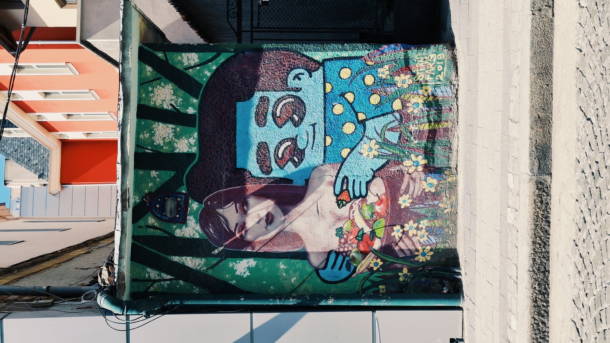 The media file shows a section of a building wall adorned with a vibrant graffiti mural. The mural depicts two figures: one appears to be a woman lying down, with her face partially visible, looking towards the viewer with a melancholic expression. She has long, dark hair and is adorned with flowers and fruit. Above her, another figure, stylized with a blue face, large eyes, and a polka-dot pattern on its clothing, is leaning over her, possibly in an embrace or protective gesture. The background of the mural is a lush green, textured with white splotches, and interspersed with dark green abstract shapes resembling branches or foliage. Small yellow flowers are scattered throughout the artwork.
To the left of the graffiti, a portion of a brightly colored building is visible, featuring orange and red sections with white-framed windows. This building appears to be part of an urban environment. To the right of the mural, the wall transitions to a textured stone surface.
The scene is brightly lit, suggesting it is daytime, likely with clear weather. The angle of the sunlight casting shadows on the mural and the building indicates the sun is at a moderate height in the sky. The presence of the building and the street-level view suggest an urban setting. The overall mood of the mural is complex, with elements of both tenderness and a touch of sadness conveyed through the figures' expressions and poses. No visible text is present in the graffiti itself, though there are some unidentifiable markings on a blue object within the mural. The media file appears to be a photograph taken in Plovdiv, Bulgaria, as indicated by the location context.