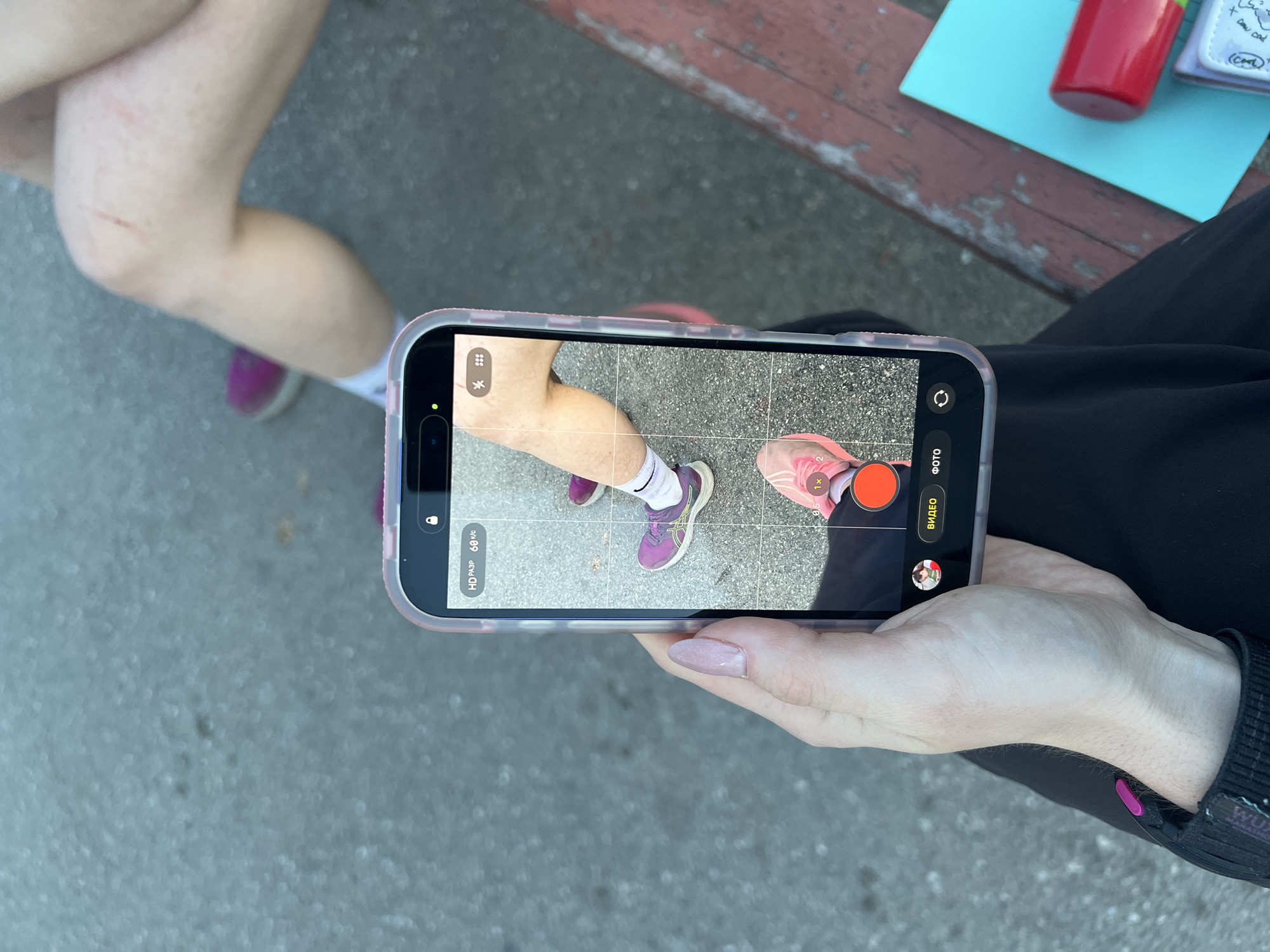 The image shows a person holding a smartphone, with the camera app open. The phone's screen displays an image of two pairs of legs and feet standing on a concrete surface. One pair of legs shows a noticeable scratch. The background includes a bench with a red container and a small notebook on it. The person holding the phone is wearing a black jacket or shirt with a visible strap bearing the text "WUX MARATHON." According to the OCR results, the notebook has the word "Cort" written on it. The location is Perm, Russia.