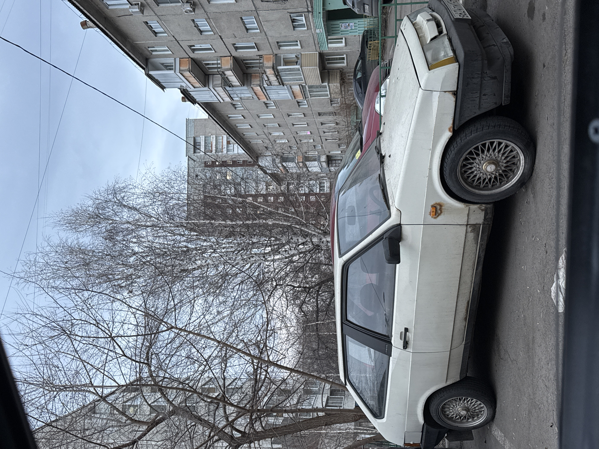 The image depicts an urban scene in Krasnoyarsk, Russia. A white Lada Samara car is parked on an asphalt road adjacent to a Soviet-era apartment building. The car appears to be in disrepair, with a damaged front bumper and some visible rust. Behind the car, there's a red vehicle partially visible. The buildings are characterized by their uniform, multi-story structure with numerous windows and balconies. A large, bare tree dominates the foreground, indicating it is likely winter or early spring. The sky is overcast, creating a muted, gray atmosphere. There are no people visible in the frame.