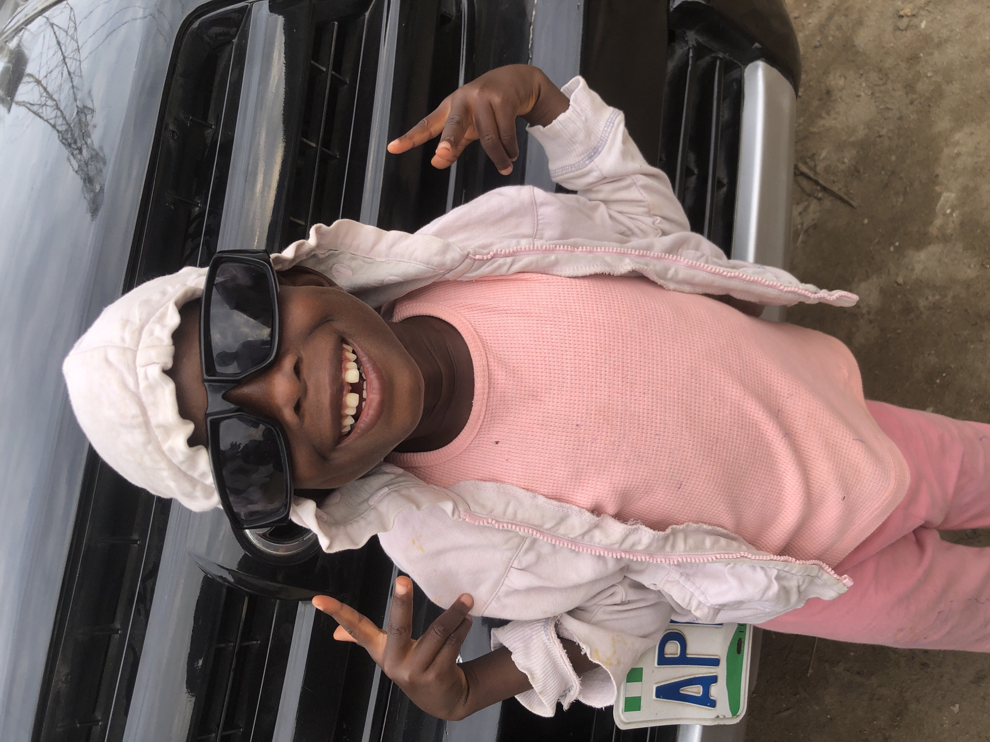 A young child, wearing a white bonnet, oversized black sunglasses, a pink t-shirt, a light pink hooded jacket, and pink pants, poses in front of a black vehicle. The child is smiling and making hand gestures with both hands, displaying two fingers up on each hand. A license plate, partially visible and reading "AP", is attached to the front of the vehicle. The background suggests an outdoor urban environment, likely in Ado, Nigeria, given the presence of the vehicle and the context of the location. The ground appears to be a mix of dirt and rough pavement.