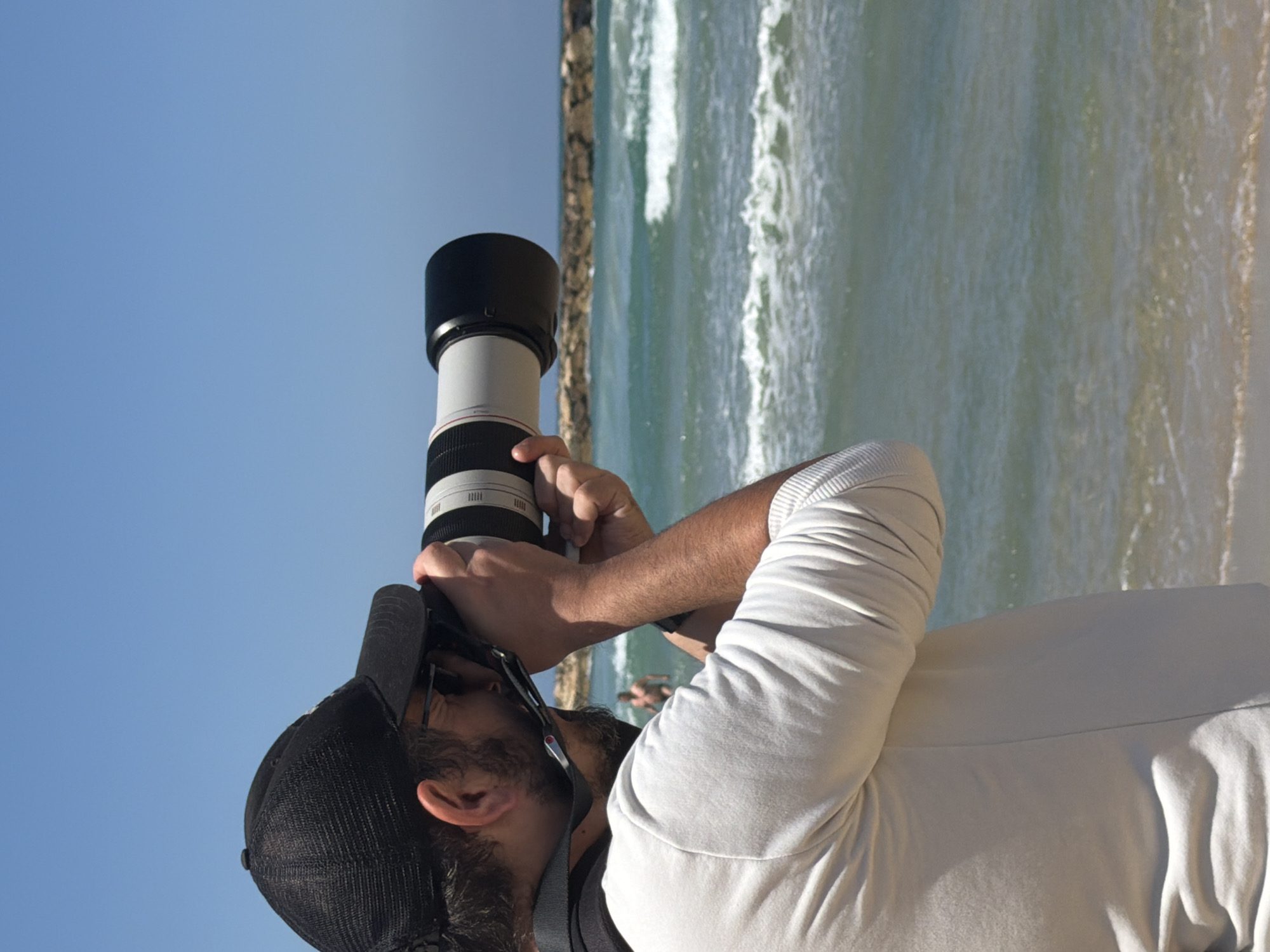 A person wearing a black cap and a light-colored, long-sleeved shirt is holding a large camera lens and appears to be taking a photograph. The background consists of a blue sky and a beach with waves. A person is seen in the distance in the water. The image was taken in Tel-Aviv, Israel.