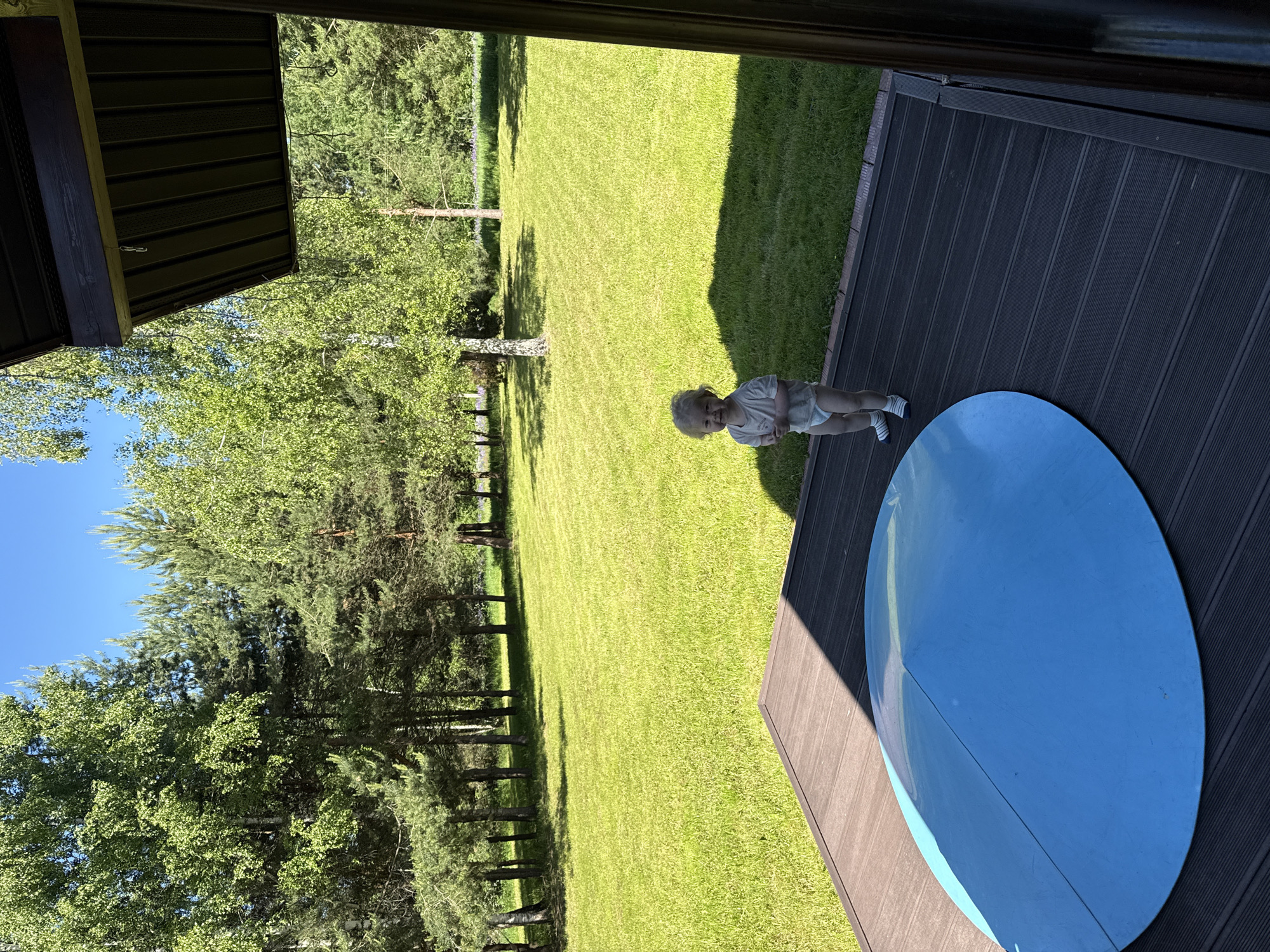 The image depicts an outdoor scene featuring a young child on a wooden deck overlooking an expansive, well-maintained lawn bordered by a variety of trees under a clear blue sky.
The location cannot be forensically confirmed as Копнино, Russia, based solely on the visual content of the image, as no specific landmarks or identifying markers are present. The flora (birch and pine trees) is typical of many forested regions.
A single child, appearing to be a toddler, stands centrally on a dark brown, slatted wooden deck. The child has light, possibly blond or very light brown, curly hair and is wearing a light-colored short-sleeved t-shirt, a white diaper, and white socks with thin blue horizontal stripes. The child faces slightly towards the viewer, exhibiting a subtle smile or happy expression, with hands clasped together at chest level.
On the deck, to the lower left of the child, rests a large, circular, convex object of bright blue color, approximately half the width of the deck. It features a distinct seam or line running across its diameter. Part of the deck, particularly on the right side and under the blue object, is cast in shadow originating from an unseen overhead structure, a portion of which (a dark brown eave or overhang with visible hooks) is visible in the upper right quadrant of the image.
Beyond the deck, a meticulously mowed, bright green lawn stretches into the midground, displaying faint horizontal mowing stripes. The lawn is bordered by a dense line of trees, including deciduous trees with light green foliage, likely birches, and various coniferous trees such as pines. In the far background, additional greenery and a patch of purple flowers are discernible. The sky above is clear and vibrant blue, indicative of fair weather. The distinct shadows suggest bright sunlight, likely during midday or early afternoon.