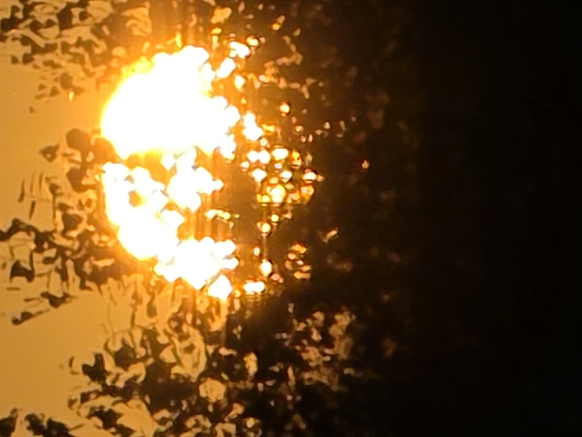 A blurred image depicts a bright light source, likely the sun, shining through a textured surface that appears to be wet or frosted. The light is diffused and appears as an intense white-yellow glow with surrounding areas of orange and dark brown. The texture on the surface creates an abstract pattern of darker shapes against the luminous background. The bottom portion of the image is almost entirely black, suggesting a shadowed area or a dark foreground. There are no identifiable people, objects, or specific environmental features from Magnitka, Russia, visible in this abstract and out-of-focus scene.