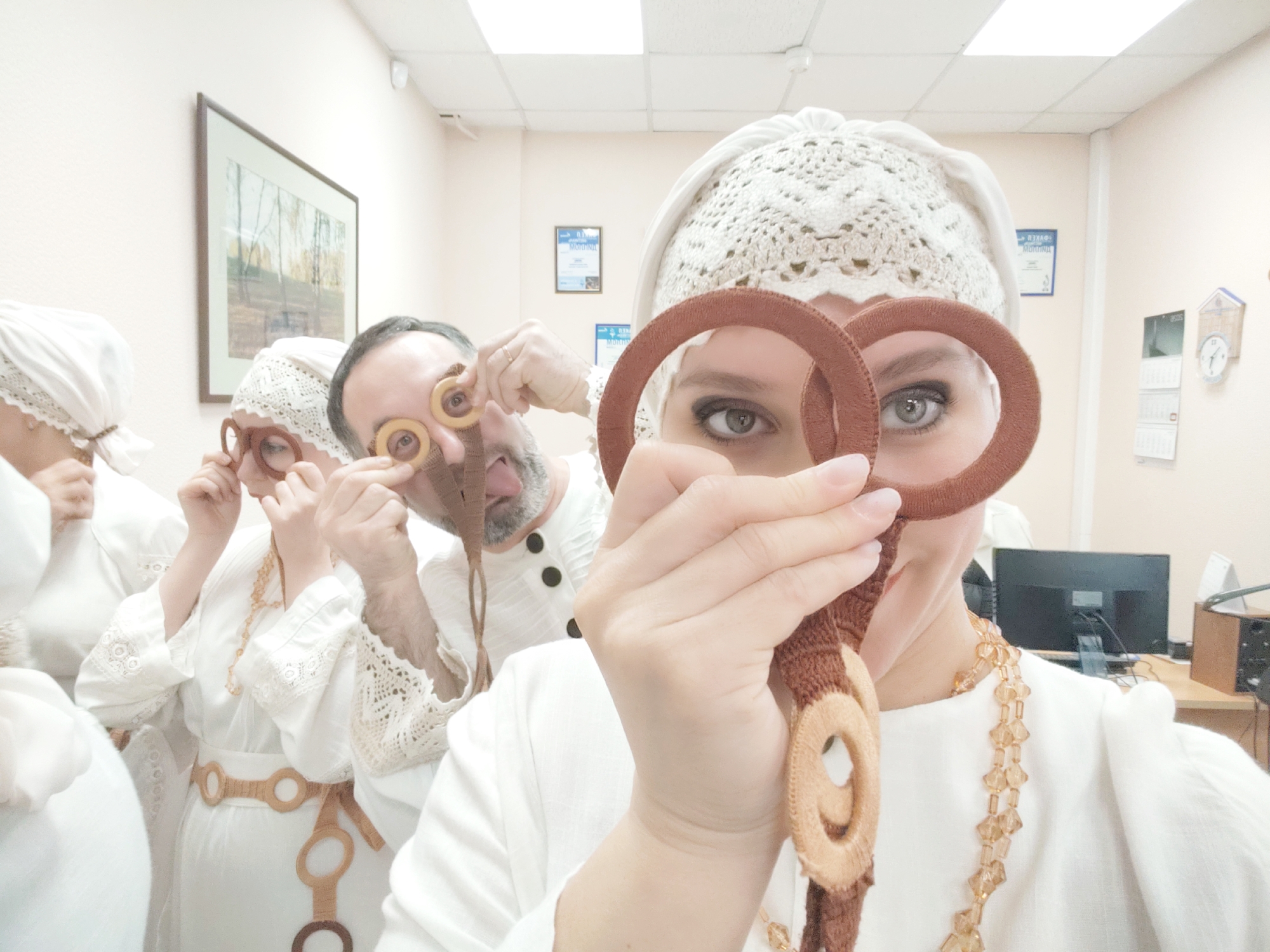A group of people, four of whom are clearly visible, are posing in an indoor environment that appears to be a room or office space. They all seem to be dressed in similar attire consisting of white clothing and white lace caps.
The person in the foreground is holding a handmade brown belt with multiple brown-and-beige rings in front of their face, obscuring part of it. The others are holding up small, circular objects to their eyes, mimicking the action of wearing eyeglasses. One man is sticking his tongue out. The belt seems to be part of their costume. The person in the front also wears a beaded necklace.
In the background, there's a framed picture on the wall and various documents, including what looks like a certificate or an award, as well as a calendar. A computer monitor and other office items can be seen on a desk.
The location is identified as Chaikovsky, Russia.