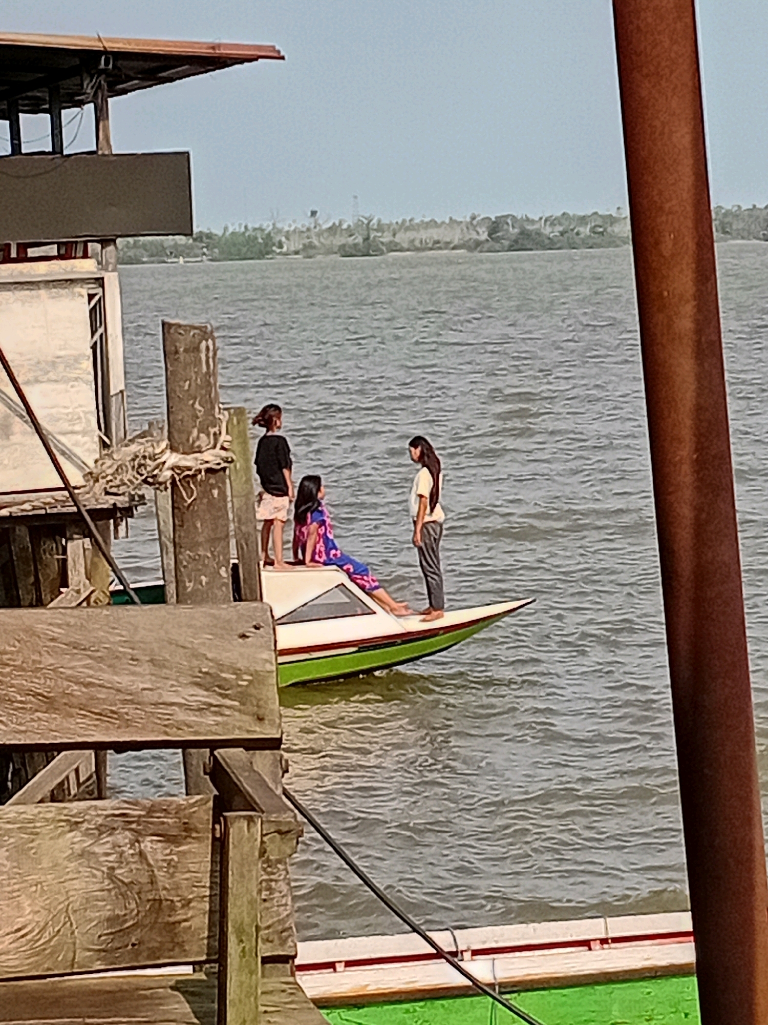 In Sungai Meriam, Indonesia, three young individuals are on a small boat. One, wearing a patterned blue and pink dress, sits on the edge of the boat, her legs extended. Another, in a black t-shirt and light-colored shorts, stands on the boat's deck. The third, dressed in a white t-shirt and grey pants, stands facing the person in the black t-shirt. The boat is on a body of water, likely a river or estuary, with a distant shoreline of trees visible. A wooden structure, possibly a pier or building, is in the foreground, partially obscuring the scene. A thick, reddish-brown pole is also prominent in the right foreground. The sky is overcast.