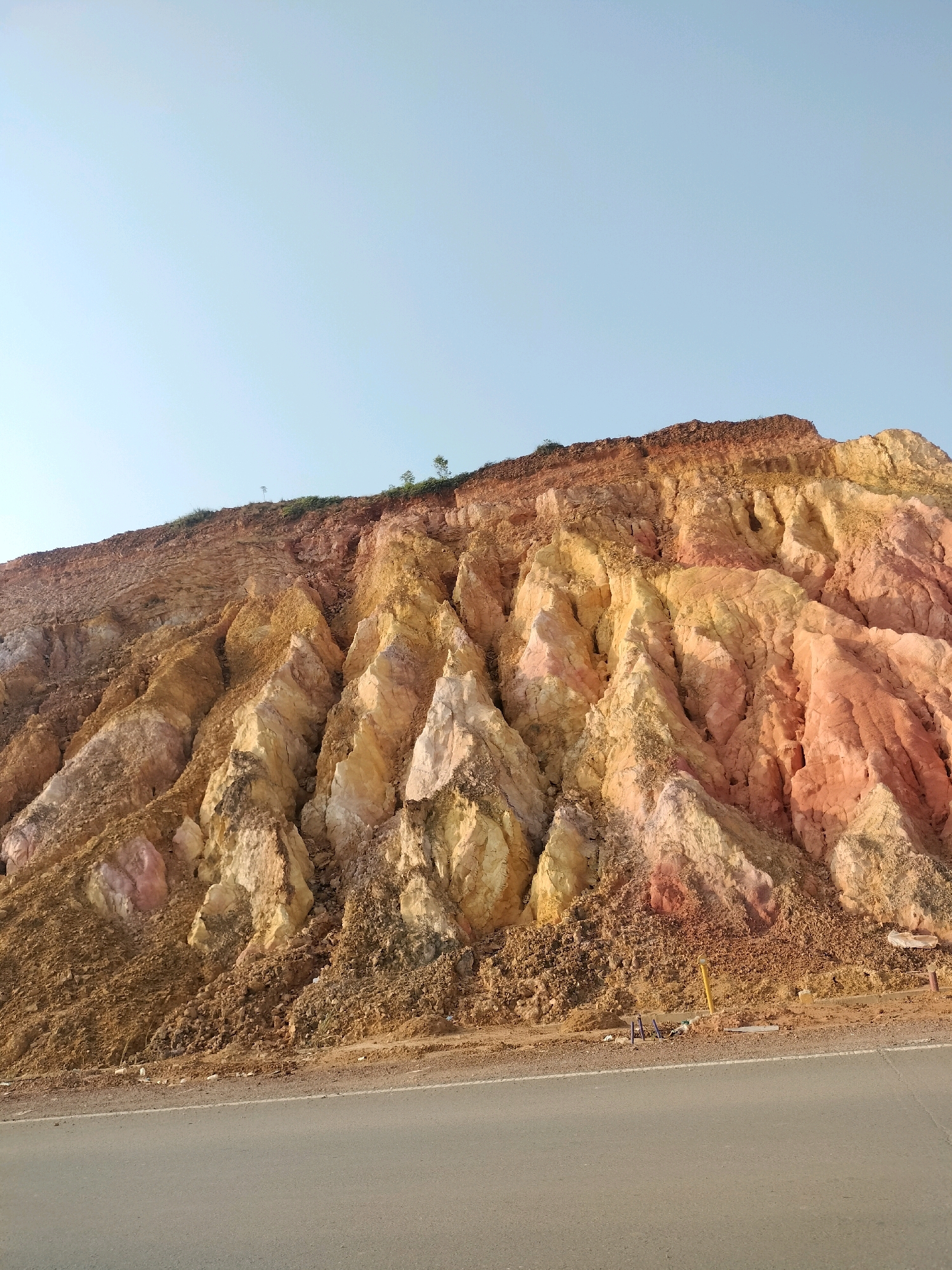 The image shows a section of a rocky hillside with layered strata of various colors, including red, yellow, and white. The hillside is steep and appears to have been eroded, creating a jagged, uneven surface with deep fissures. The top of the hill has some sparse vegetation and a utility pole. In the foreground, a paved road runs horizontally across the bottom of the image. The sky is clear and light blue. The scene depicts a natural landscape with road infrastructure. Based on its appearance and the context of Batam, Indonesia, this could be a road cut through a hilly area, possibly near a construction site or a natural geological formation.