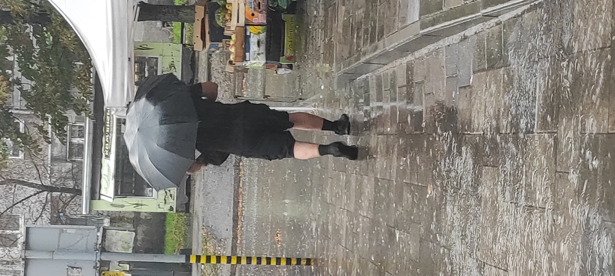 A person is walking in the rain, holding a black umbrella over their head. The individual is wearing black shorts, a black t-shirt, black socks, and dark shoes. The pavement is wet and reflective, with visible rain falling. To the right of the person, there are stacked boxes containing fruits. In the background, there is a small kiosk with a green facade and a white tent structure. A yellow and black striped pole is visible to the left of the frame. The overall scene suggests an outdoor market or street setting in the city of Kamianske, Ukraine, during inclement weather.
