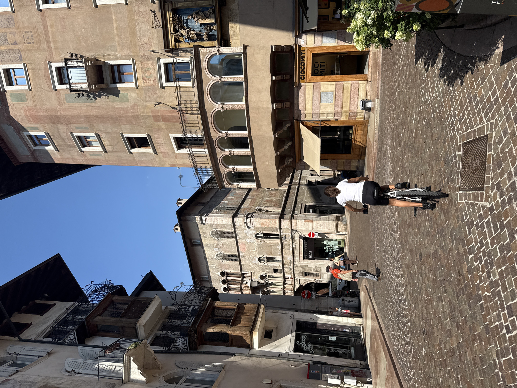 The image depicts a cobblestone street in the city of Trento, Italy, on a sunny day. In the foreground, a woman on a bicycle is riding away from the camera. She is wearing a white t-shirt and black shorts. The street is lined with historic buildings. On the right side of the image, a building features an ornate facade with arched windows and a shop entrance with the signage "CIOCMITI". On the left side, another building has balconies with intricate wrought-iron railings. In the background, other pedestrians can be seen walking on the street. The sky is clear blue.