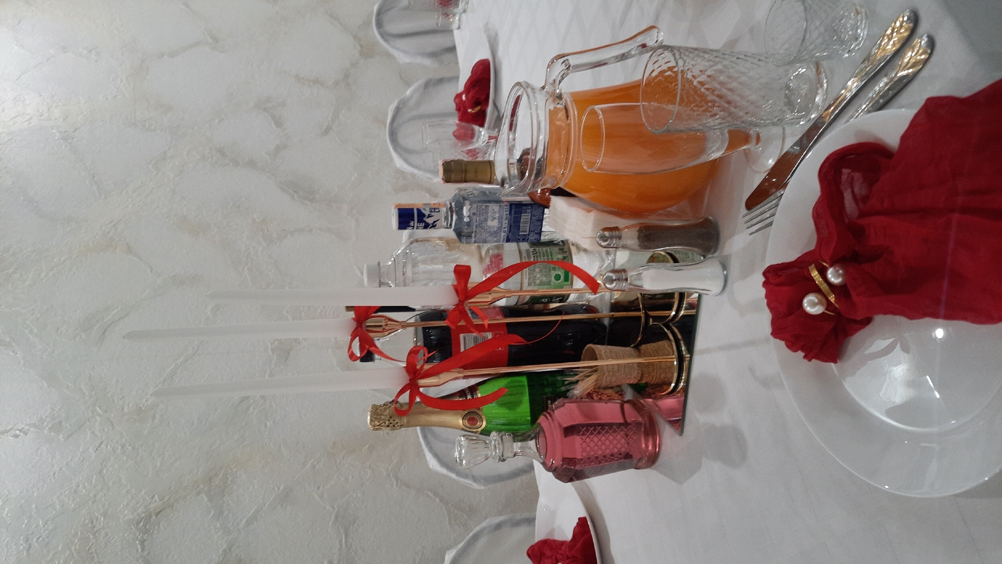 The image depicts a table setting at what appears to be a formal event in the village of Zhodzishki, within the Zhodzishki village council of Belarus. The table is covered with a white tablecloth. A centerpiece features three tall, white candles in a gold candelabra adorned with red ribbon. Surrounding the candelabra are various bottles, including a green bottle of alcohol, a dark bottle of soda, a clear bottle of water, and a blue and white bottle of spirits. A decorative pink glass decanter with a stopper is also present. To the right of the centerpiece, a glass pitcher filled with orange liquid, likely juice, is visible along with two drinking glasses, one partially obscured by the pitcher. In the foreground, a white plate holds a folded red napkin secured with two pearl-like rings. Silverware, including a knife and fork, is laid out on the right side of the plate. In the background, several white chairs are visible, suggesting a dining area. The wall behind the table has a textured, light-colored finish.