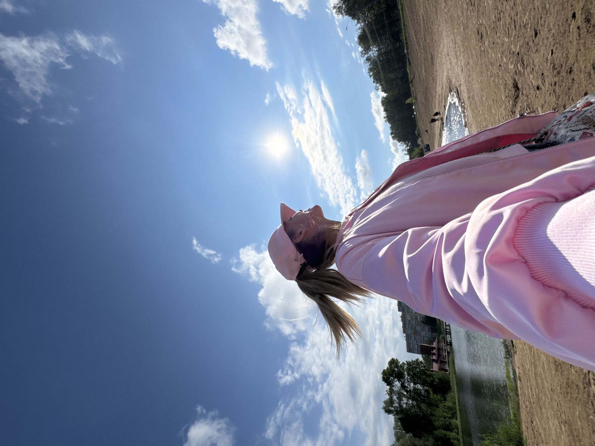 The image shows a woman in a pink outfit, possibly a jacket and cap, looking up at the sky. The sky is bright blue with scattered white clouds, and the sun is visible. In the background, there is a landscape that includes a body of water, trees, and what appears to be a sandy or dirt ground. Buildings can be seen in the distance, potentially indicating an urban or suburban environment. Based on the landscape features, it is likely that the location is in Saint Petersburg, Russia, which has numerous bodies of water, trees, and open spaces, including parks with bodies of water, surrounded by urban infrastructure. The overall impression is a sunny day with the woman enjoying the view of the sky and landscape.