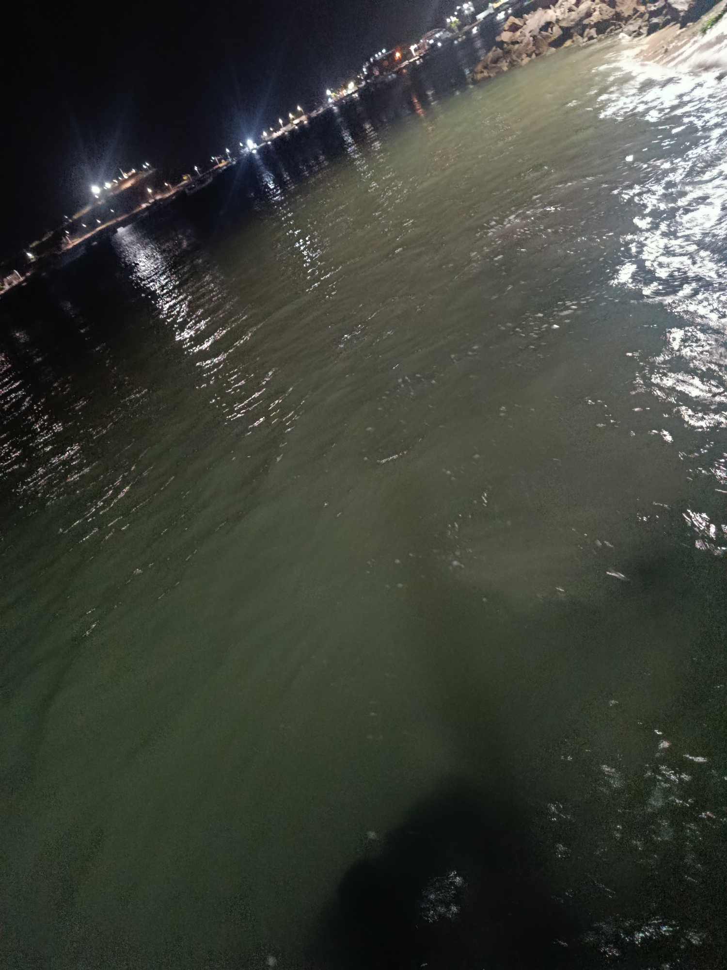The image depicts a night scene in what appears to be Juan Griego, Venezuela, showing water with rippling surfaces reflecting light. The water is dark, likely due to the night, and the reflections indicate some illumination along the coastline. There are some street lights, and a few faint lights suggesting the presence of buildings or structures further inland. The bottom of the image is darker, possibly indicating the presence of a shadow or something submerged in the water near the viewer's vantage point.