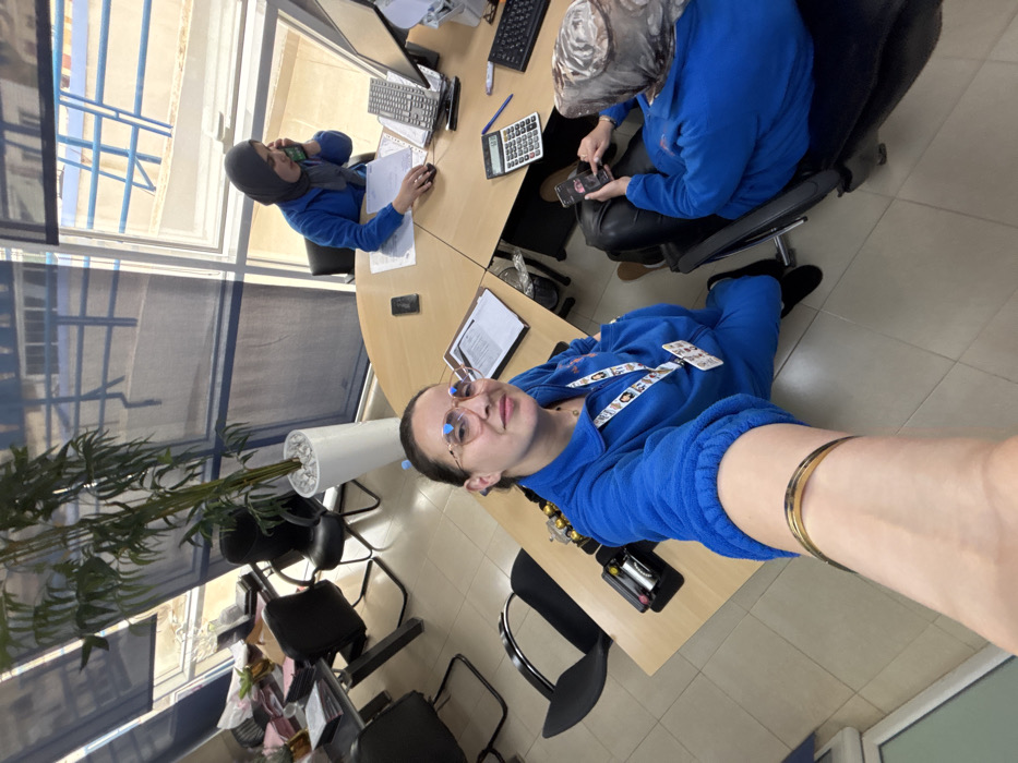 This image depicts an indoor office environment in Meknes, Morocco, captured from an elevated perspective as a selfie. Three women are present, all wearing blue, fleece-like long-sleeved tops.
In the foreground, a woman with dark hair and gold-rimmed glasses is seen from the chest up, taking the photo. She wears an ID badge on a patterned lanyard and a gold-colored bracelet on her left wrist. Her expression is a faint smile.
To her right, further into the mid-ground, a second woman is seated at a light-colored modular desk. She wears a grey head covering and is holding a green corded phone to her right ear. Her right hand operates a computer mouse, while a computer monitor, keyboard, and various papers are arranged on the desk in front of her.
To the lower right, partially obscured, a third woman is seated in a black office chair, wearing a patterned light brown/grey head covering. She is looking down and actively interacting with a black smartphone held in both hands. Her legs are crossed.
The office features multiple light-colored wooden desks, black office chairs, and a light-tiled floor. Large windows line the back wall, partially covered by blue roller blinds. Through the uncovered sections of the windows, a light blue railing and exterior structures are visible, suggesting an upper floor. A tall, artificial bamboo-like plant in a white pot with white stones is positioned near the windows. Desk accessories include a black organizer with pens/brushes, smartphones, a calculator, and various documents.