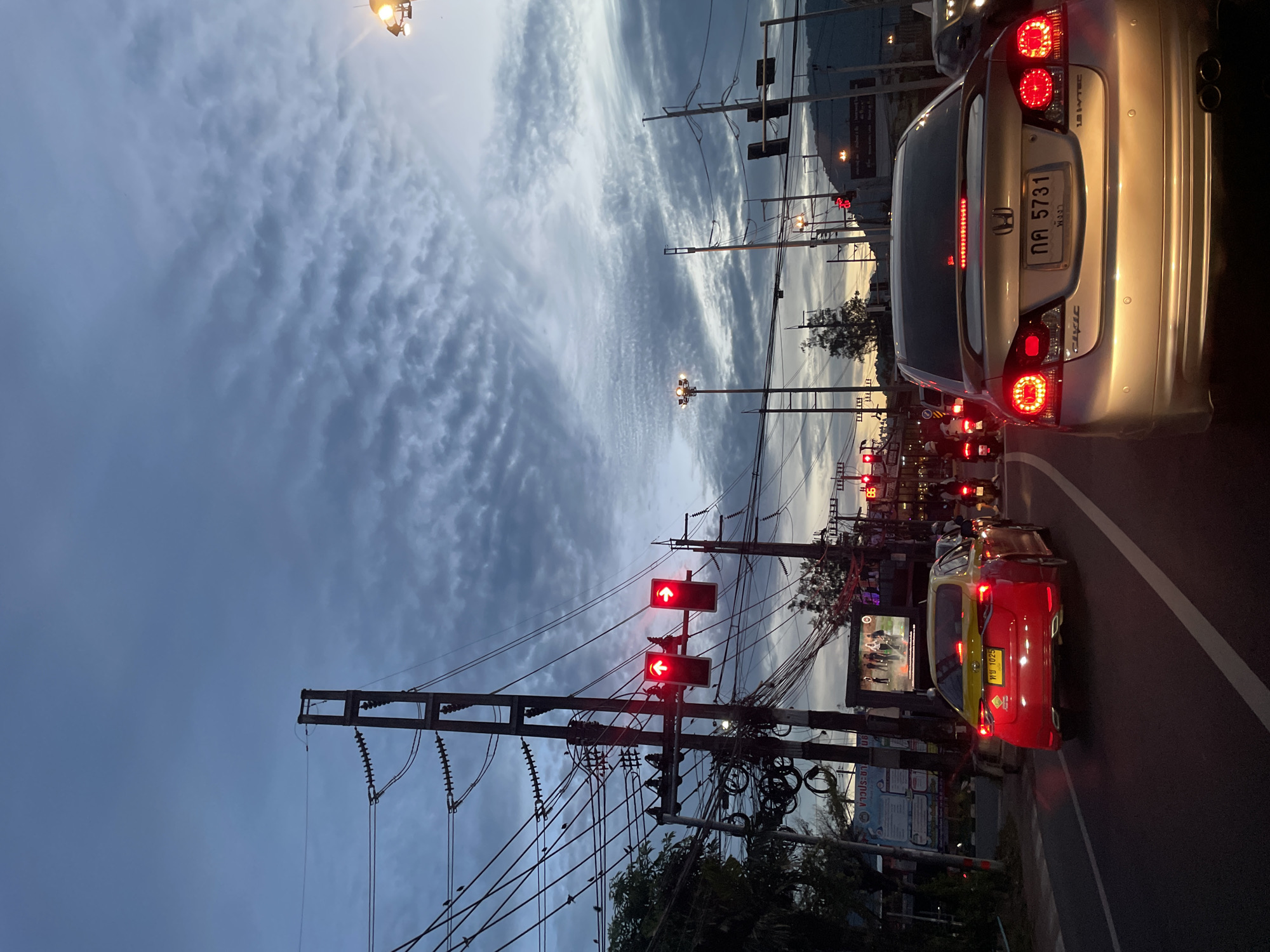 The image shows a street scene at dusk in an urban environment. The sky is overcast with textured clouds. In the foreground, the rear of a silver Honda Civic is prominently visible, its brake lights illuminated. The car has a license plate reading "กต 5731" in Thai script. To the left, a red and yellow taxi is stopped at a traffic light, with its license plate reading "พษ 1025". Traffic lights are visible, with at least two showing red signals. Utility poles with numerous wires are present throughout the scene. In the background, there are indications of buildings, streetlights, and a distant hillside. The overall impression is of a city street during evening rush hour, with vehicles stopped at an intersection.