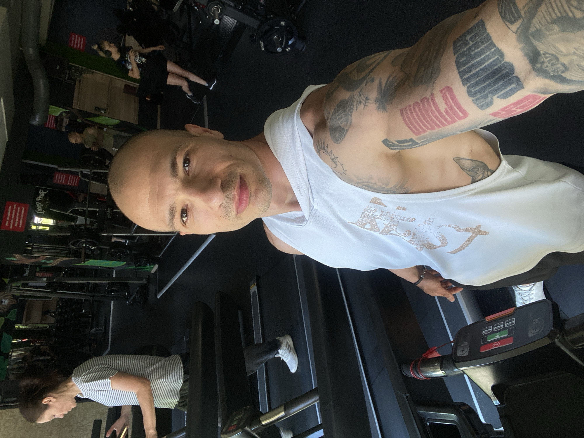 A man is taking a selfie at a gym in Pleven, Bulgaria. He is bald, wearing a white tank top, and has tattoos on his right arm. He is standing on a treadmill. There are other people exercising in the background. Gym equipment is visible, including weights and weight machines.