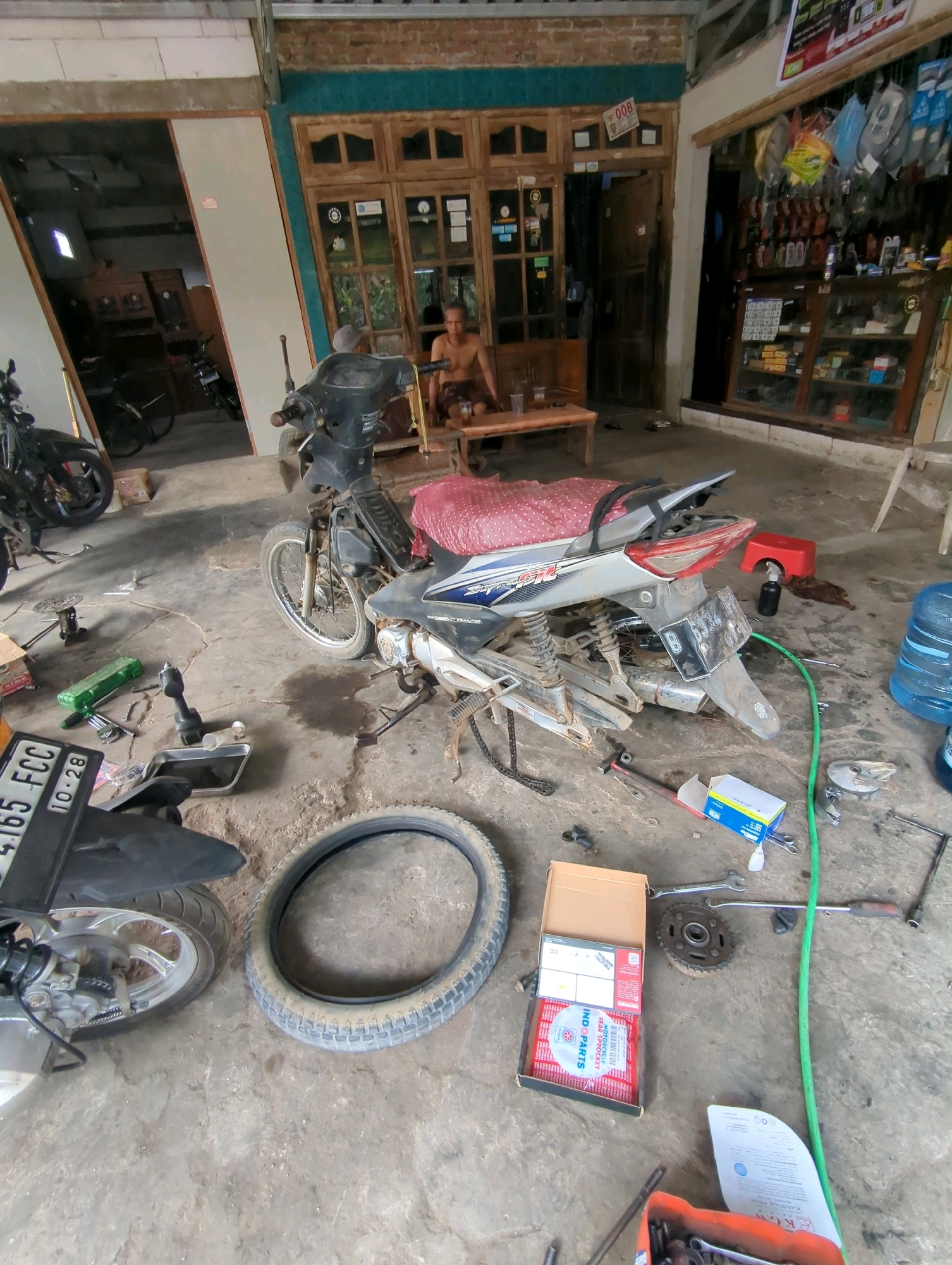 The image depicts a cluttered motorcycle repair shop in Kedungdowo, Indonesia. A partially disassembled motorcycle, silver with blue accents and a pink seat cover, is the central focus. Tools and spare parts are scattered across the concrete floor, including a detached tire, wrenches, a drill, and boxes of motorcycle components. To the left, another motorcycle is visible, partially obscured. In the background, a man sits on a wooden bench inside a dimly lit building with wooden doors and windows. The shop appears to be attached to a residential dwelling, with shelves displaying various items in the background. The overall impression is one of a functional, albeit messy, workshop.