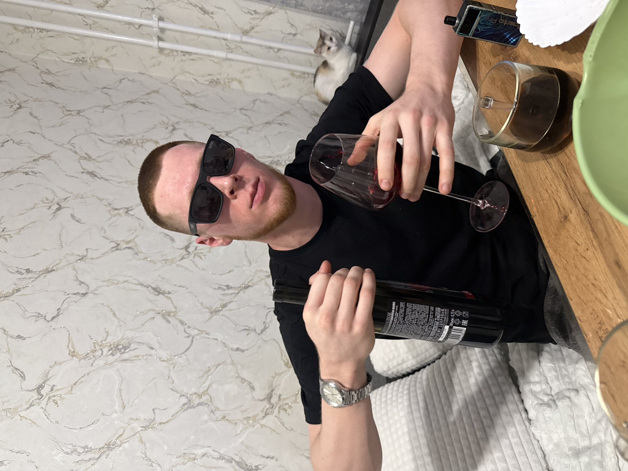 A man wearing sunglasses and a wristwatch is pouring red wine from a black bottle into a wine glass. He's sitting at a wooden table with a smartphone, a small container, and a green bowl on it. There's a white cat in the background, and the scene appears to be indoors, with a white patterned wall and a white corduroy-like sofa.