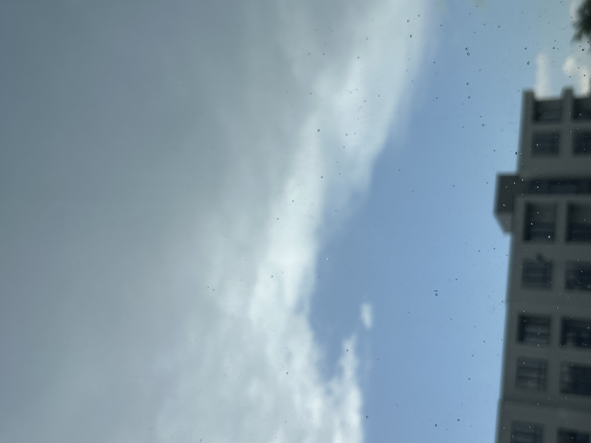 The image depicts a view of the sky and a building, possibly taken through a window or windshield. The sky is partly cloudy with both dark, overcast areas and patches of blue. A tall, gray building with multiple windows is visible on the right side of the frame, though its details are somewhat blurred. The presence of water droplets on the glass suggests it may have been raining. There's no information in the image to identify this location in Plovdiv, Bulgaria.