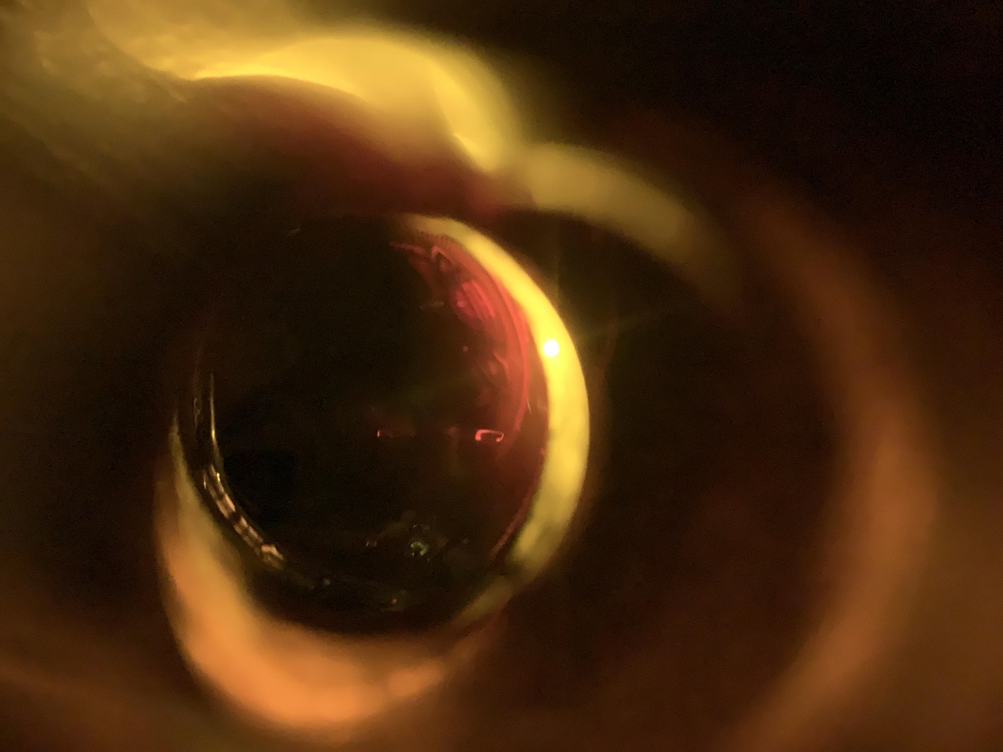 The image shows a blurred, close-up view of a round object, possibly a glass or lens, with a reflective surface. The object is illuminated with a warm, golden light, creating a halo effect around its edges. There are reddish and dark tones visible within the object's interior. The background is dark and out of focus, with hints of brown and golden hues. The location is not discernible, and there is no indication that this is New Stadium, Nigeria.