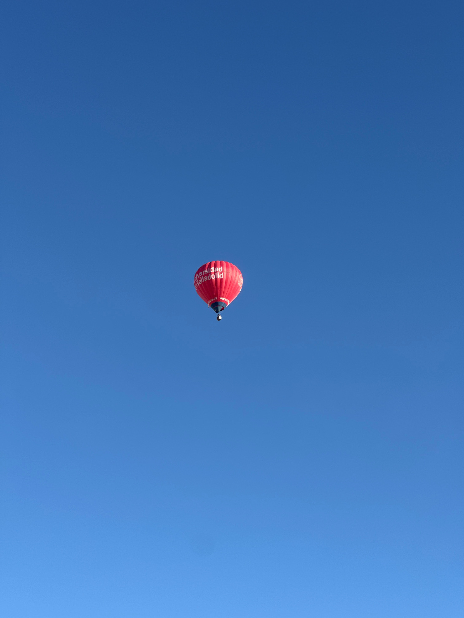A single red hot air balloon is visible in the sky over Arroyo de la Encomienda, Spain. The balloon is positioned in the center-right of the frame and appears to be ascending. It is adorned with white lettering that reads "Universidad de Valladolid" and "www.uva.es". The sky is a clear, bright blue, with no clouds present. The bottom of the balloon features a dark gondola. The image was captured on a sunny day, as indicated by the clear sky.