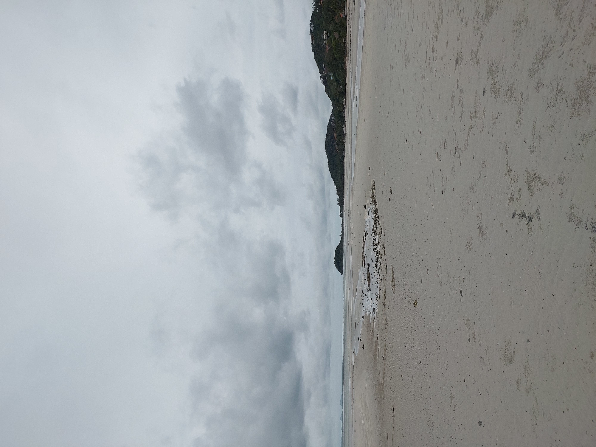 A wide-angle, eye-level shot captures a desolate, overcast beach scene in Ko Samui District, Thailand. The foreground is dominated by a vast expanse of wet, pale sand, marked by subtle ripples and scattered small dark pebbles or shells, suggesting the recent ebb of the tide. In the mid-ground, a shallow body of water reflects the muted sky, with more scattered rocks and debris visible along its edge. Beyond the water, a dark, conical island rises from the sea, its peak covered in dense green vegetation. Further to the right, two more hills, also covered in lush foliage, are visible, dotted with numerous white structures, indicating human habitation. The sky is filled with heavy, grey clouds, creating a somber and tranquil atmosphere. The image depicts a natural coastal landscape under a cloudy sky.