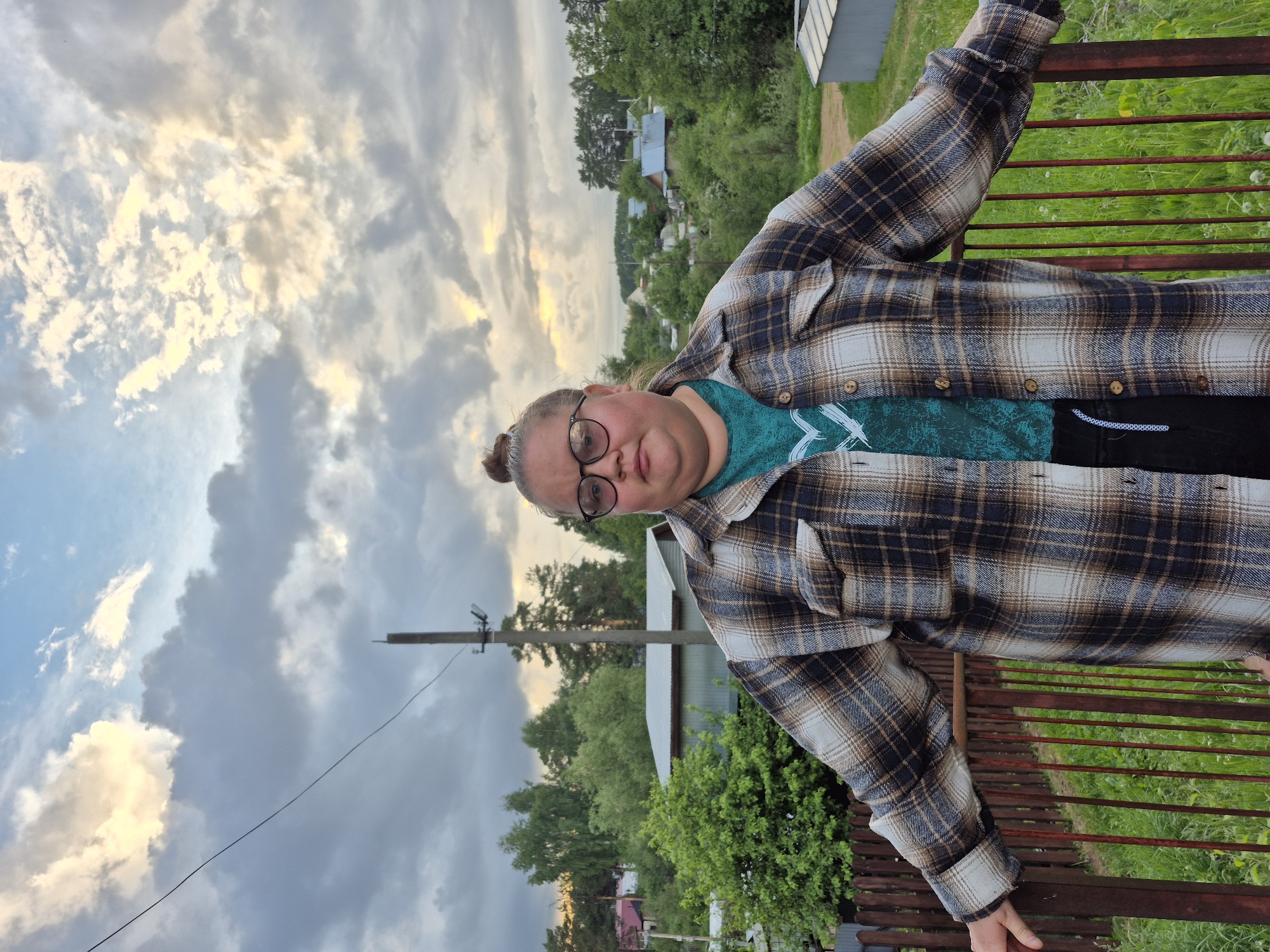 An outdoor portrait captures a person with light hair tied in a bun and wearing black-rimmed glasses, a dark green t-shirt with a white graphic design, and an open plaid shirt (brown, black, and cream) over it. The individual is looking directly at the camera with a neutral expression, leaning casually on a rusty, dark red metal fence. Their arms are resting on the top bar of the fence.
Behind the person, the scene reveals a semi-rural landscape under a dramatic, cloudy sky. In the foreground, green grass and dense foliage are visible through the fence bars. The mid-ground features a wooden utility pole with overhead wires, prominent against the sky. Further back, scattered houses with grey metal roofs are nestled among abundant green trees and foliage, suggesting a village or countryside setting. A dirt path can be seen winding through the distant landscape.
The sky is filled with a dynamic mix of light and dark grey clouds, with patches of golden-yellow and bright white light breaking through, indicative of either sunrise or sunset. The overall atmosphere is tranquil and natural. No visible text is present in the image. The setting is consistent with a location like Chaikovsky, Russia.