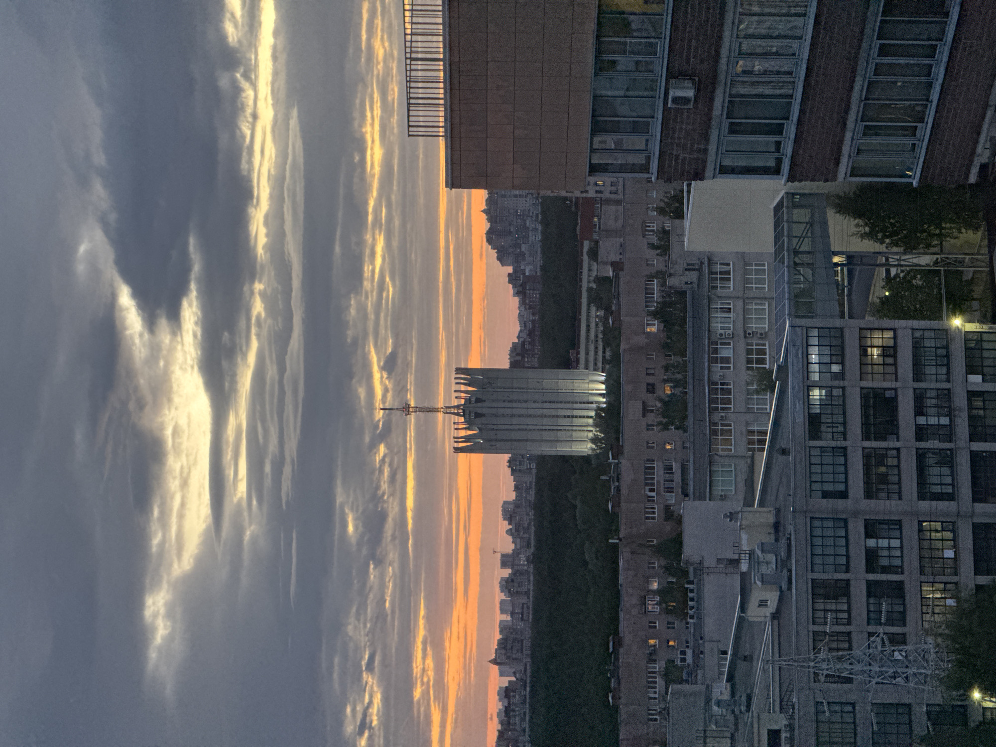 The image depicts an urban scene at twilight. A tall, modern building with a communication tower is prominent in the foreground, silhouetted against a sunset with orange and grey clouds. Below the building is a line of dense trees, suggesting a park or green space. Beyond the trees are more buildings, indicating a city skyline. To the right, several multi-story apartment buildings are visible, some with lights on in the windows. In the foreground on the right, a utility structure (likely a power transformer) is present. The image appears to have been taken from an elevated position, potentially a high-rise building. The orientation of the image is vertical.