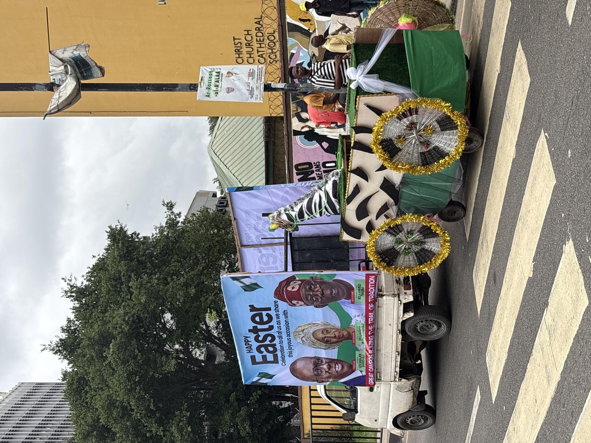 The image shows a decorated truck driving on a zebra crossing on a street in Lagos, Nigeria. The truck is adorned with a "Happy Easter" banner featuring the images of Bola Ahmed Tinubu, Dr. Claudiana Ibijoke Sanwo-Olu and Gov Babajide Sanwo-Olu. The truck also has decorations including zebra-patterned fabric and umbrellas. The background includes buildings such as Christ Church Cathedral School. People are visible on the truck and standing beside it. There are also posters visible on a wall next to the truck.
