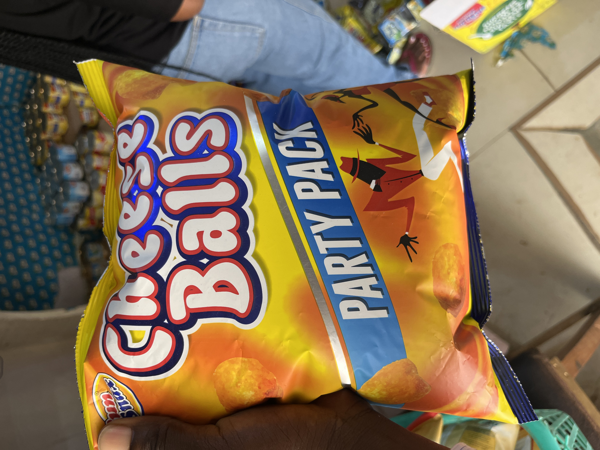 In Bukuru, Nigeria, a close-up shot captures a hand holding a yellow and orange bag of "Cheese Balls" snack. The bag prominently displays the product name in large, white, red, and blue lettering, with "Party Pack" written in white on a blue banner below. The background reveals shelves stocked with various canned goods and other packaged items, suggesting a retail environment. A person wearing denim jeans and a black top is partially visible on the right side of the frame, with their lower body in view. The lighting is bright, and the focus is on the snack bag.