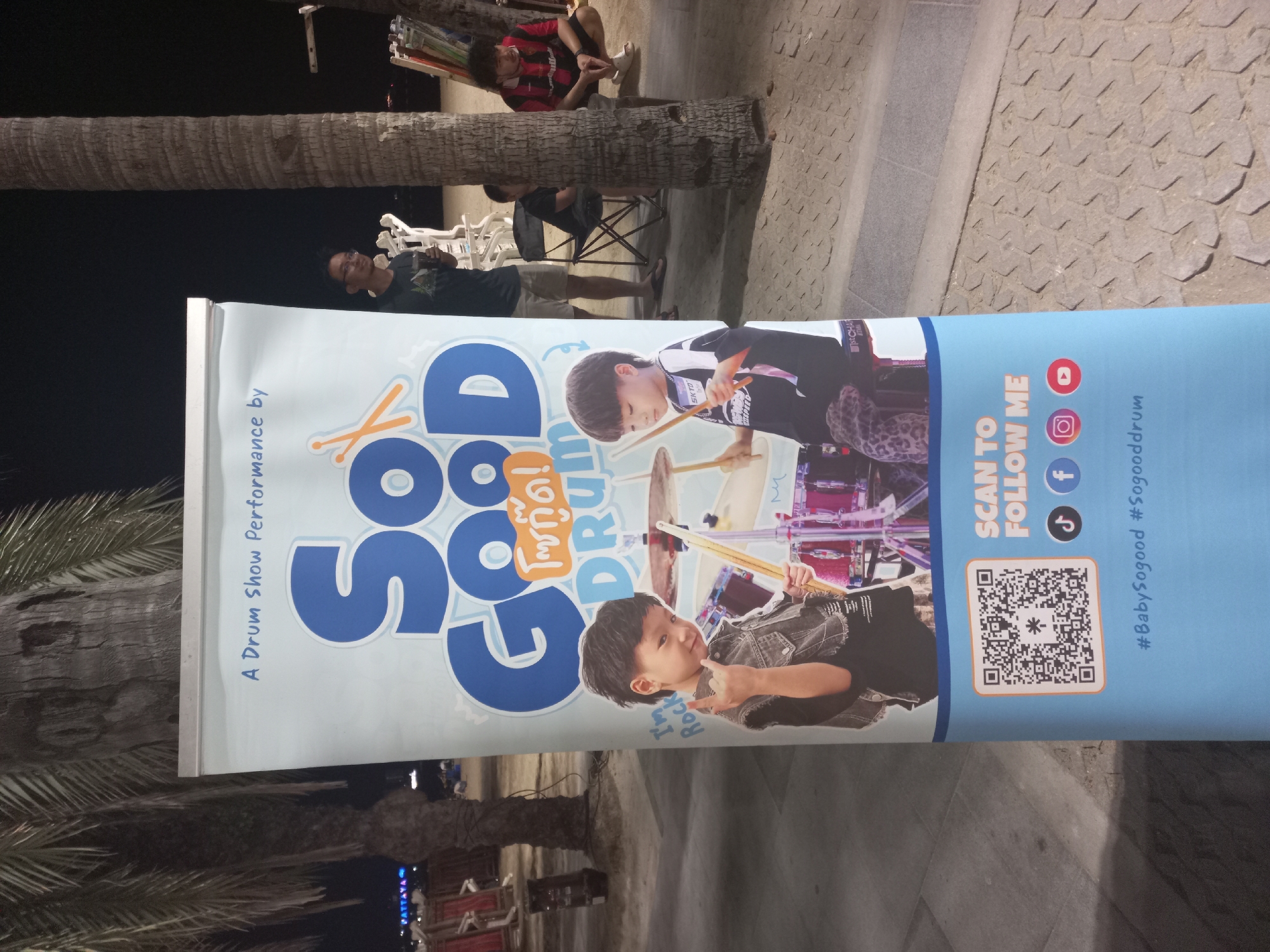 The image shows a banner advertising "Sogood Drum" next to a palm tree in Pattaya City, Thailand. The banner features images of two young drummers and includes social media links and a QR code. In the background, people are sitting and walking near the beach. The environment appears to be outdoors in the evening.