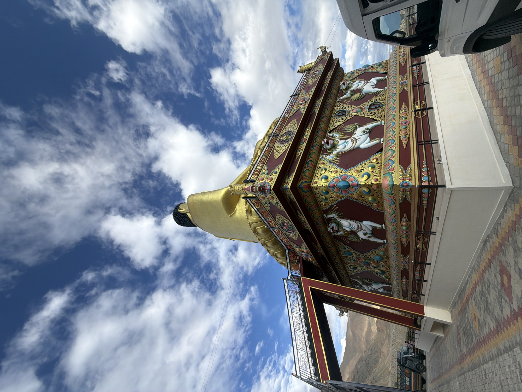 The image depicts a large, golden Buddha statue situated in Leh, India. The statue is adorned with intricate, colorful decorations featuring mythical creatures. In the foreground, a white van is parked, and a truck is visible in the background. The scene is set against a backdrop of mountains and a cloudy blue sky. The ground is paved with interlocking bricks in a geometric pattern.