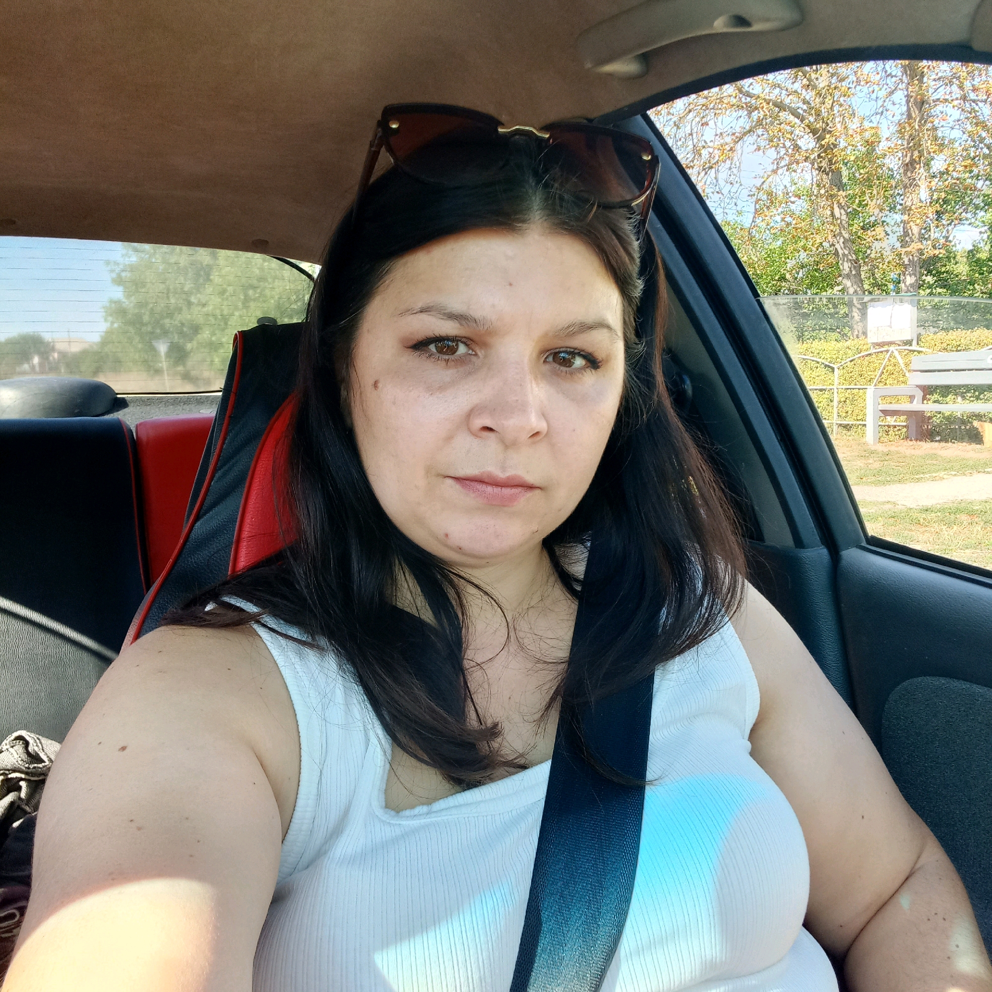 In Klyntsi, Ukraine, a woman with dark hair and brown eyes is taking a selfie inside a car. She is wearing a white tank top and sunglasses are resting on top of her head. The interior of the car is visible, with black and red seat upholstery and a seatbelt across her chest. Sunlight casts shadows on her. Outside the car window, trees and a park bench can be seen.