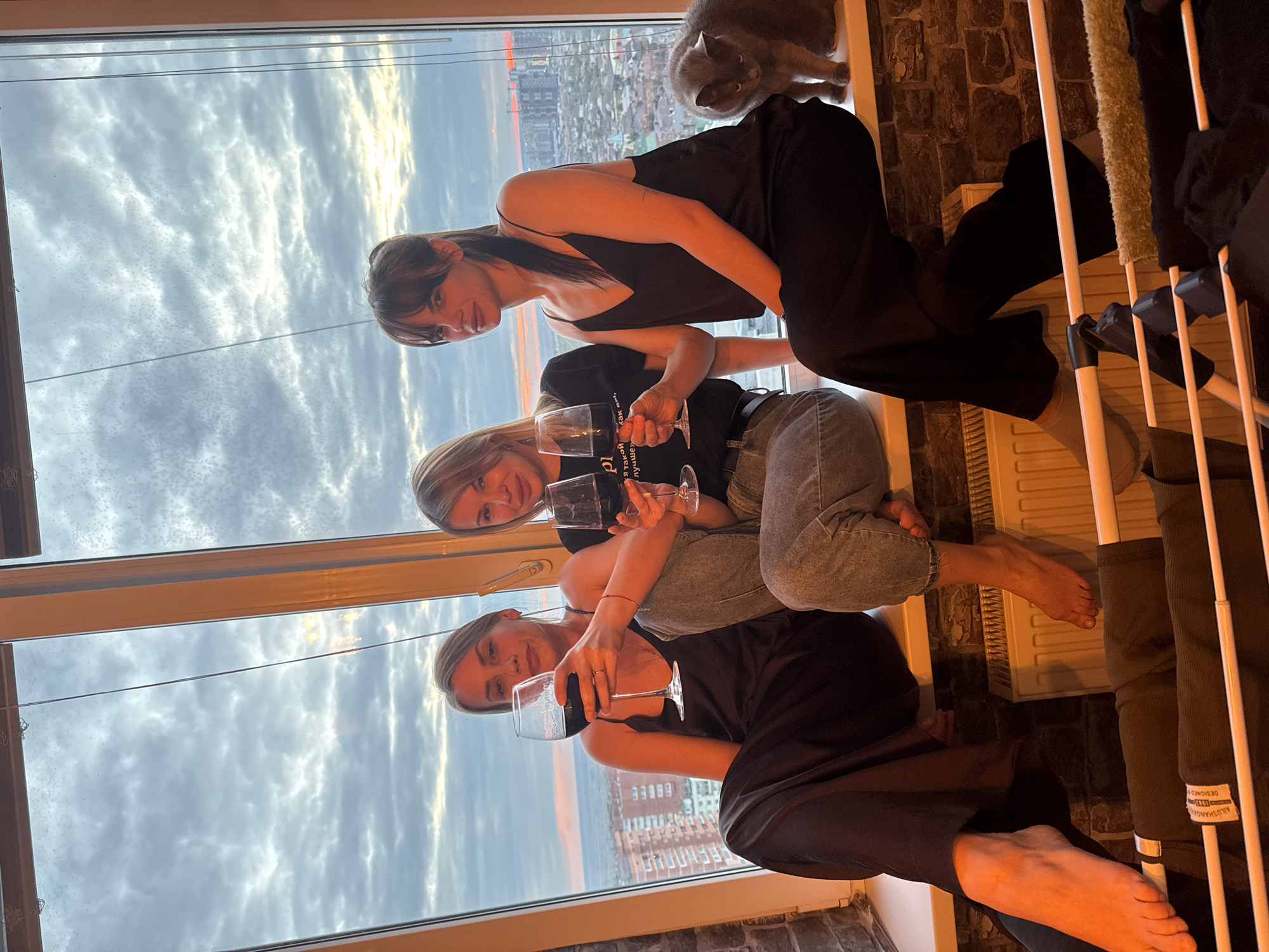 Three women are seated on a window ledge, holding wine glasses. Behind them is a window displaying a cityscape at sunset. The skyline indicates the city of Rostov-on-Don, Russia. A grey cat is also perched on the windowsill. A clothes rack is situated beside the women.
