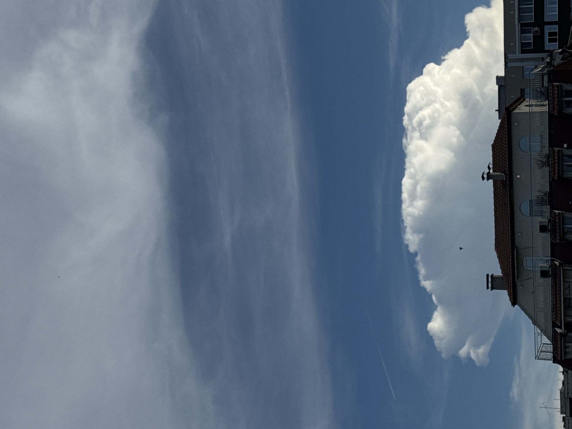 A vertical, medium shot captures a clear blue sky with scattered white clouds and a distant building in Plovdiv, Bulgaria. The sky occupies the majority of the frame, featuring wispy cirrus clouds high up and a large cumulus cloud formation lower down. A faint contrail from an airplane is visible on the left side of the sky. In the lower portion of the image, the top of a building with a terracotta-tiled roof and dormer windows is visible. Chimneys protrude from the roof. Another building with darker facade and windows is situated behind the first building. The overall scene depicts a sunny day with a dynamic sky above an urban landscape.