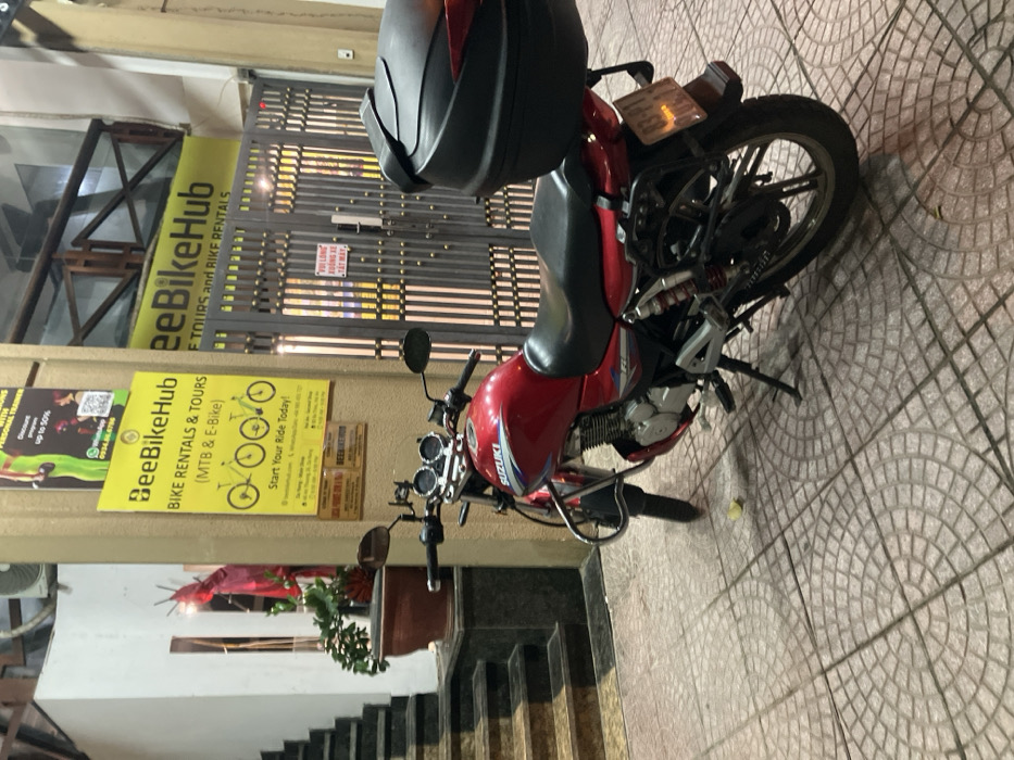 An outdoor, nighttime scene in Ngũ Hành Sơn Ward, Vietnam, depicts a red Suzuki motorcycle parked on a tiled sidewalk in front of a building entrance. The motorcycle, equipped with a black seat, chrome crash bars, and a black hard-shell top case, faces left with its kickstand deployed. Its rear license plate, partially visible, begins with "43-P1". The sidewalk features square gray tiles with circular relief patterns. To the left of the motorcycle, a dark-colored staircase ascends next to a large brown potted plant containing green foliage and red decorative items. To the right of the motorcycle, a light-colored building pillar displays two vertical yellow banners. The lower banner advertises "BeeBikeHub BIKE RENTALS & TOURS (MTB & E-BIKE)" with bicycle graphics. The upper banner is a "PERSONAL TRAINER" advertisement with a WhatsApp number and QR code. Adjacent to the pillar is a dark gray metal gate, partially revealing an illuminated interior. A white sign with red text on the gate reads "VUI LÒNG XUỐNG XE TẮT MÁY" (Please get off the bike and turn off the engine). Above the gate, another yellow "BeeBikeHub" sign is visible. No individuals are present in the scene.