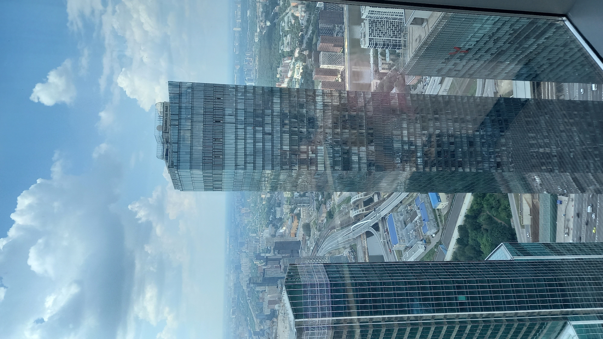 The image is a high-angle, vertical shot of a modern city skyline. In the foreground, two glass-covered skyscrapers dominate the frame. The skyscraper on the right is taller and more prominent, featuring a facade with a grid pattern of windows and reflective surfaces. The skyscraper on the left is shorter and wider, also covered in glass panels.
Below the skyscrapers, a complex network of railway tracks and bridges is visible, weaving through the urban landscape. A river or large body of water can be seen in the middle ground, reflecting the sky and buildings. Beyond the immediate area, a sprawling cityscape stretches to the horizon, characterized by numerous buildings of varying heights and architectural styles. The sky is partially filled with white, fluffy clouds against a pale blue backdrop. The overall scene depicts a dense, modern urban environment.