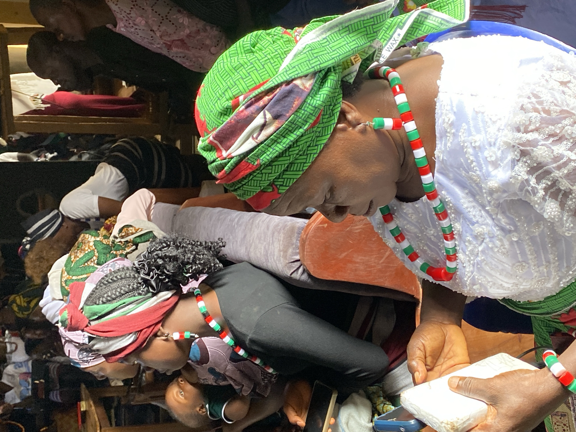A scene depicts multiple individuals within a seemingly crowded and possibly indoor environment. Predominantly, the focus is on a few people. One person, wearing a green and patterned headwrap along with a necklace and bracelet made of red, green, and white beads, is holding a white rectangular object. This person is also wearing a white lace top. Another person nearby is wearing a similar beaded necklace and bracelet. The clothing style and adornments might indicate a cultural or traditional setting.
The environment appears to be busy and possibly retail-oriented, as there are elements in the background that could be shelves or display areas with various items. The lighting suggests an indoor setting, and the overall composition and orientation of the image is slightly unusual, as the image is rotated sideways.
Given the location in Durumi, Nigeria, it's reasonable to infer the scene could be a market, gathering, or perhaps a religious ceremony. Without additional context, the specific action or interaction remains unclear, but the emphasis on certain individuals and their attire provides a focal point within the overall visual narrative.