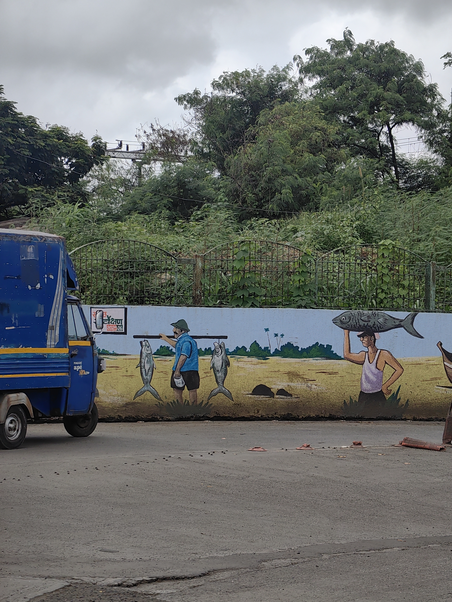 The image depicts a street scene in Mira-Bhayander, India, under a cloudy sky. On the left, a blue auto-rickshaw is parked, showing signs of wear and tear, with "Piaggio Ape Xtra" branding visible. A mural adorns a wall behind the auto-rickshaw. The mural features two men, one wearing a blue shirt and a hat, carrying fish on a pole over his shoulder, and another man wearing a white tank top, balancing a fish on his head. Both men are depicted with cartoonish features, and they are standing in a painted landscape. Behind the mural, a green metal fence encloses an area with lush green trees and foliage. The foreground shows a paved road with some debris scattered on it, including small red cone-like objects and orange plastic debris. The overall impression is of a somewhat weathered but vibrant urban environment.