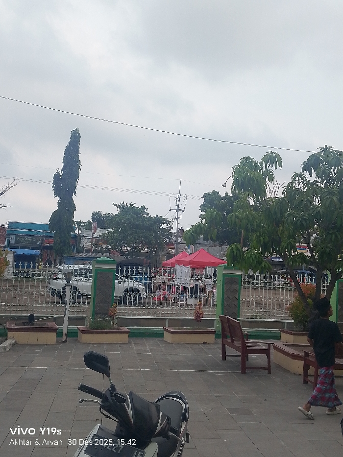 The image depicts an outdoor public area in Jagapura Kidul, Indonesia, under an overcast sky at approximately 15:42 on December 30, 2025. The foreground is a paved surface composed of light gray square tiles. A white and black scooter is parked on the left side of this paved area, facing slightly left. To the right, dark brown wooden benches are visible, along with planter boxes featuring green and yellow trim, containing small plants.
A white metal fence with decorative elements and pillars accented with green and dark gray separates the paved area from a street or market. Beyond this fence, in the midground, multiple red canopy tents are erected, indicating a market or temporary gathering. Several vehicles, including at least one white pickup truck, are parked or moving near these tents. Indistinct human figures are visible within and around the tent area, suggesting activity. Buildings with various colored facades, including blue and light brown, line the background street. Utility poles and overhead power lines are visible against the sky.
On the far right of the paved area, a single individual is walking towards the right. This person is attired in a dark-colored top and a red and blue plaid sarong-like lower garment. Trees are present throughout the scene; a tall, slender tree stands on the left background, while a tree with dense green foliage is prominent on the mid-right. The sky is uniformly light gray, indicating cloudy weather conditions.
