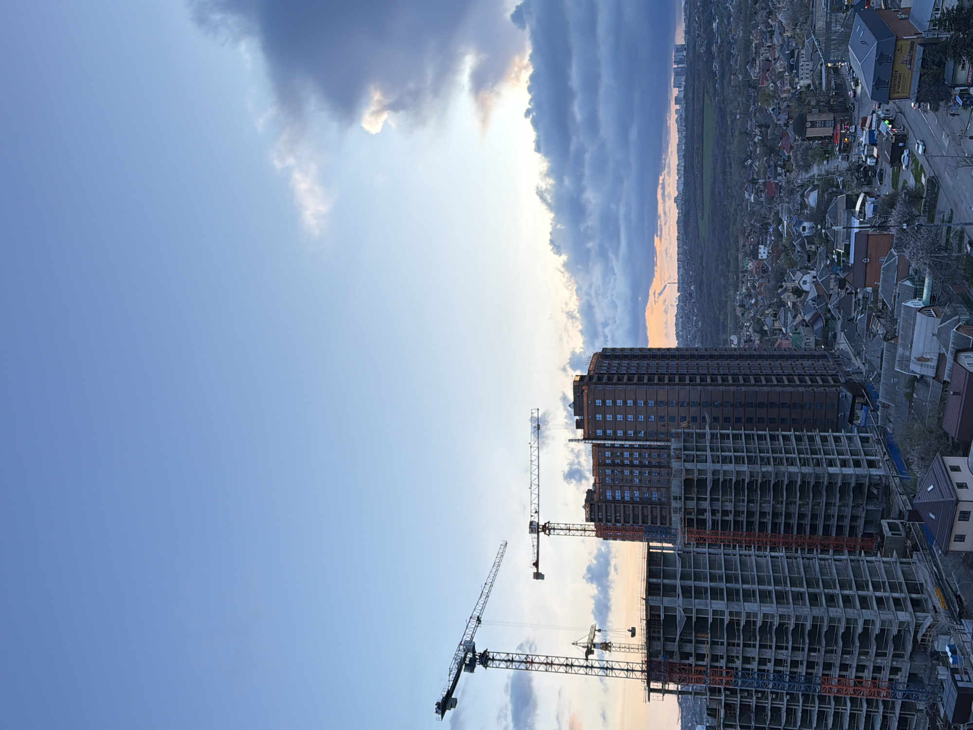 The image depicts a high-angle view of Rostov-on-Don, Russia. The foreground shows the construction site of a multi-story building complex, with several cranes in operation. Behind the construction site, there is a completed apartment building. In the background, a residential area with various houses and buildings extends towards the horizon, with a green area separating it from the cityscape in the far distance. The sky is partly cloudy, with shades of blue and grey. Traffic is visible on the roads.
