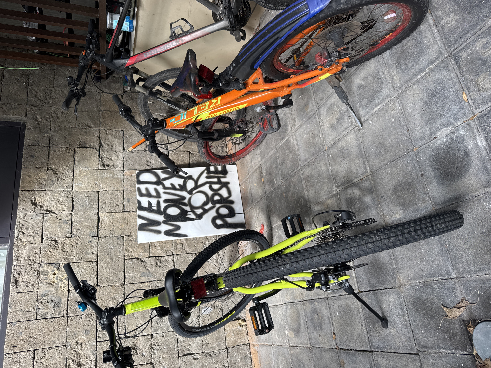 The scene is outdoors, likely in Kuta Selatan, Indonesia, based on visual cues typical of the region's built environment. Three bicycles are present. One is black and red, another is orange and black with red rims, and the third is chartreuse green. A white sign leaning against a wall displays black spray-painted text "NEED MONEY FOR PORSCHE." The ground is paved with gray stone tiles. There are also some leaves.