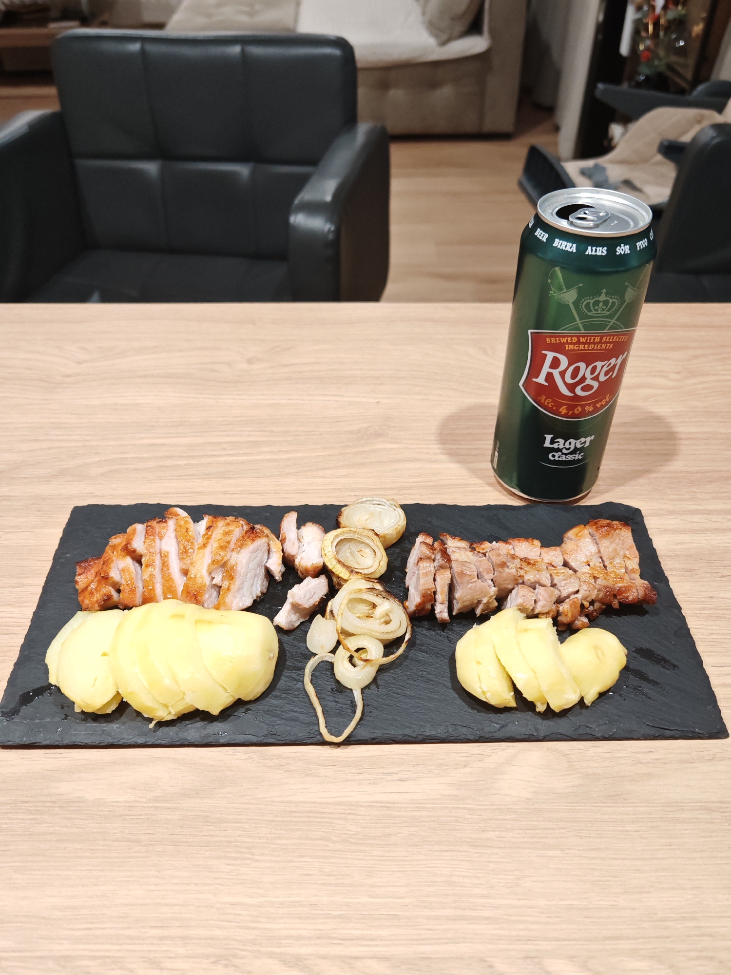 The image shows a slate platter with sliced meat, sliced potatoes, and cooked onions arranged on a wooden table. A can of Roger Lager Classic beer stands to the right of the platter. In the background, a black leather armchair is visible, along with a sofa and what appears to be a Christmas tree decorated with red ornaments. The lighting suggests an indoor setting, likely a home or restaurant. The specific location in Plovdiv, Bulgaria, cannot be determined from the provided image.