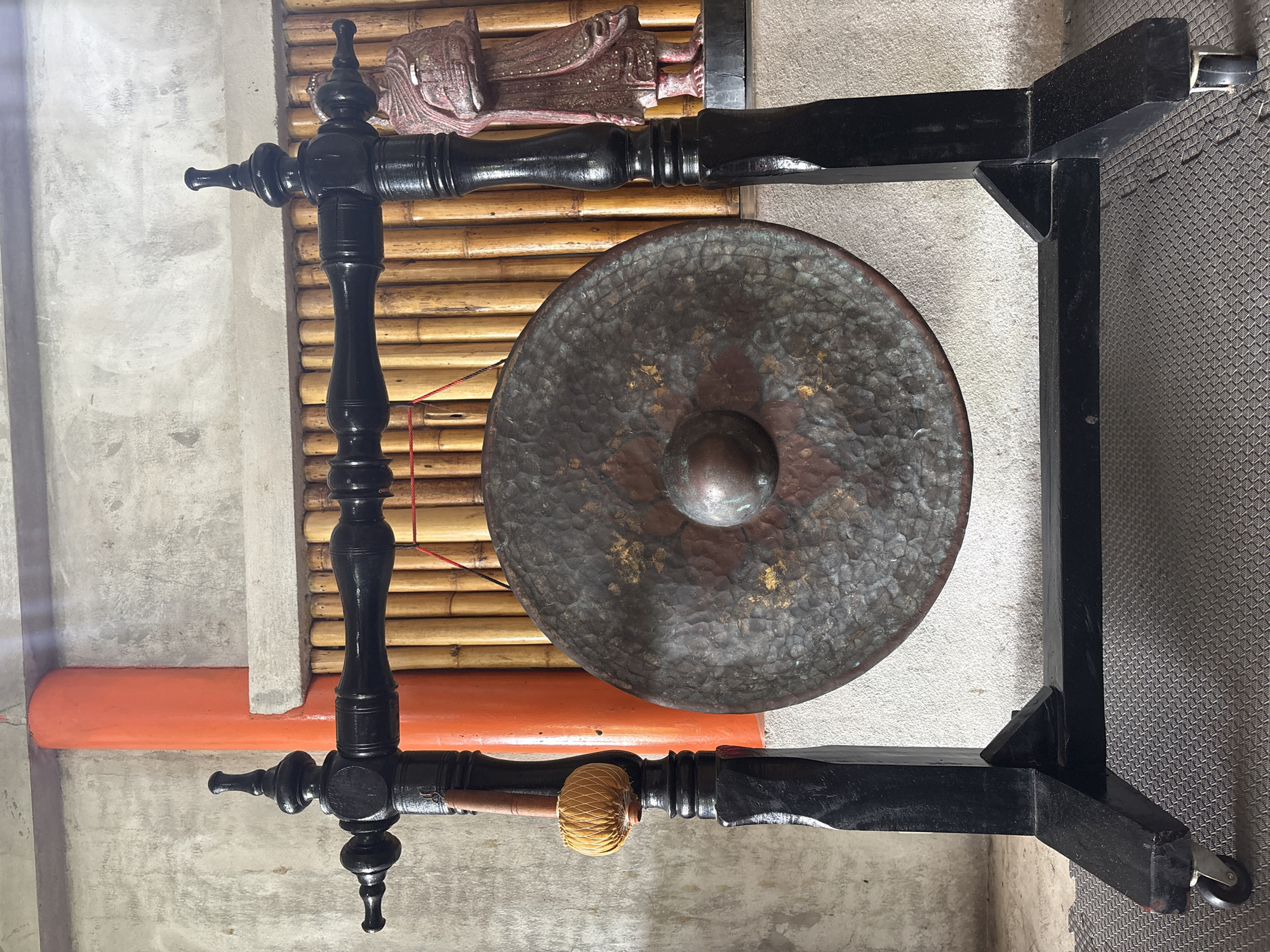 The image features a large, round gong suspended within a black wooden frame. The gong is textured and appears to be made of dark metal, with a raised center. The wooden frame has ornate turned posts and a simple, rectangular structure. A mallet with a woven head rests on one of the frame's horizontal supports.
The background includes a wall covered in bamboo slats and a concrete wall with a painted orange stripe. The flooring is a grey textured surface.
The location is Rawai, Thailand.