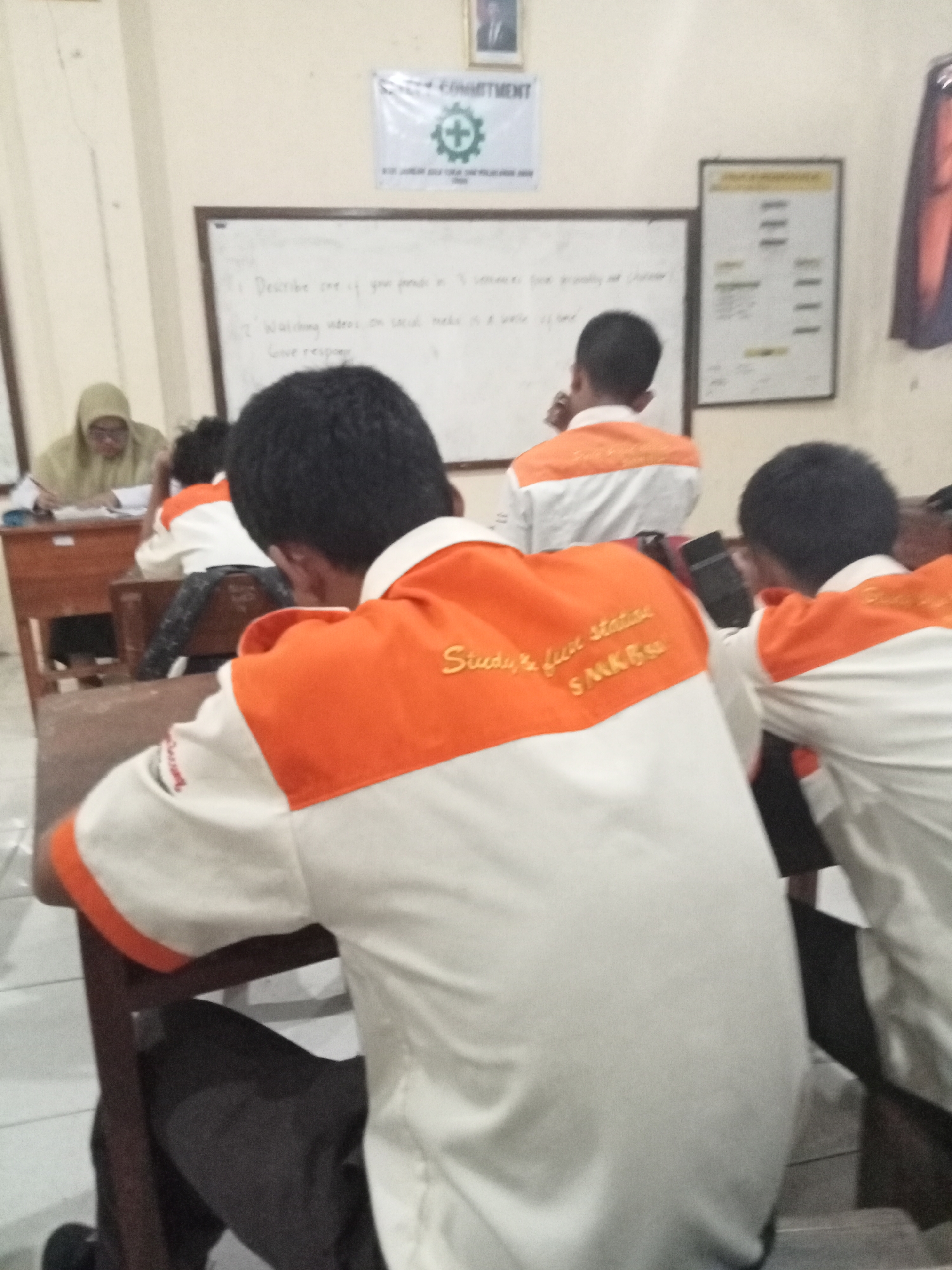 The image depicts a classroom scene at SMKN 1 Secang, located in Secang, Indonesia. In the foreground, two students are seated at desks, their backs to the camera, wearing white shirts with orange accents. The student on the left has the words "Study & Save status" and "SMKBS" embroidered in gold on their shirt. A teacher, wearing a light brown hijab, is seated at a desk on the left side of the image, attending to papers. A whiteboard with handwritten text and questions is visible on the wall behind the students. A safety banner with a green cross symbol and text is also displayed. The environment suggests a typical Indonesian vocational school classroom.