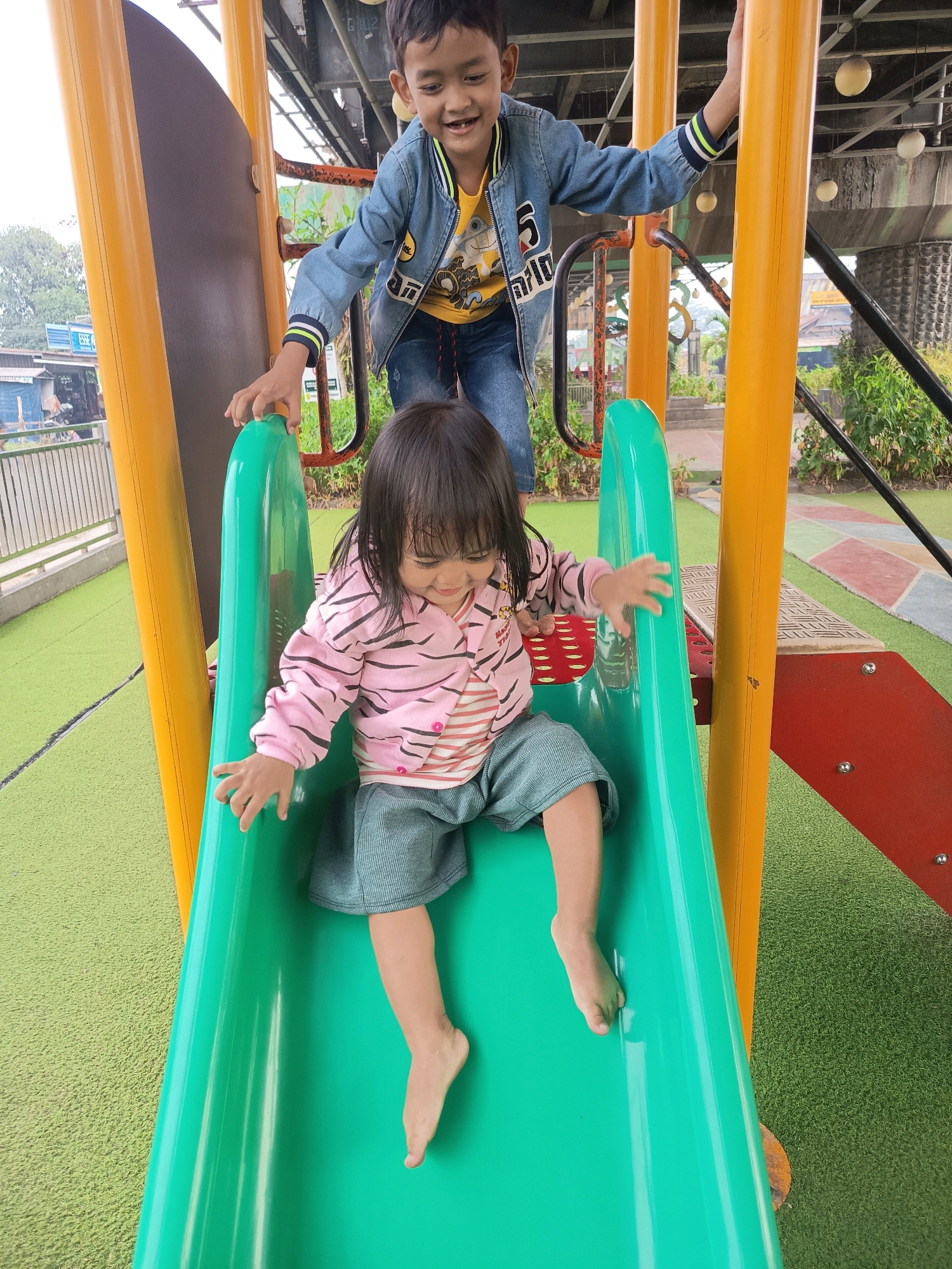 Two children are on a playground slide. A younger child, a girl with dark hair, is sliding down the green plastic slide, wearing a pink and black striped long-sleeved shirt, and blue shorts. Her legs are bare. An older child, a boy with dark hair, is standing on the platform above the slide, wearing a blue denim jacket over a yellow shirt and blue jeans. He is smiling. The playground features yellow and orange metal poles, with a red section of the slide structure visible to the right. The ground is covered in green artificial turf. In the background, a fence and some buildings are visible, along with some trees and foliage. The context of Banjarmasin, Indonesia, suggests this is likely an outdoor recreational area in the city.