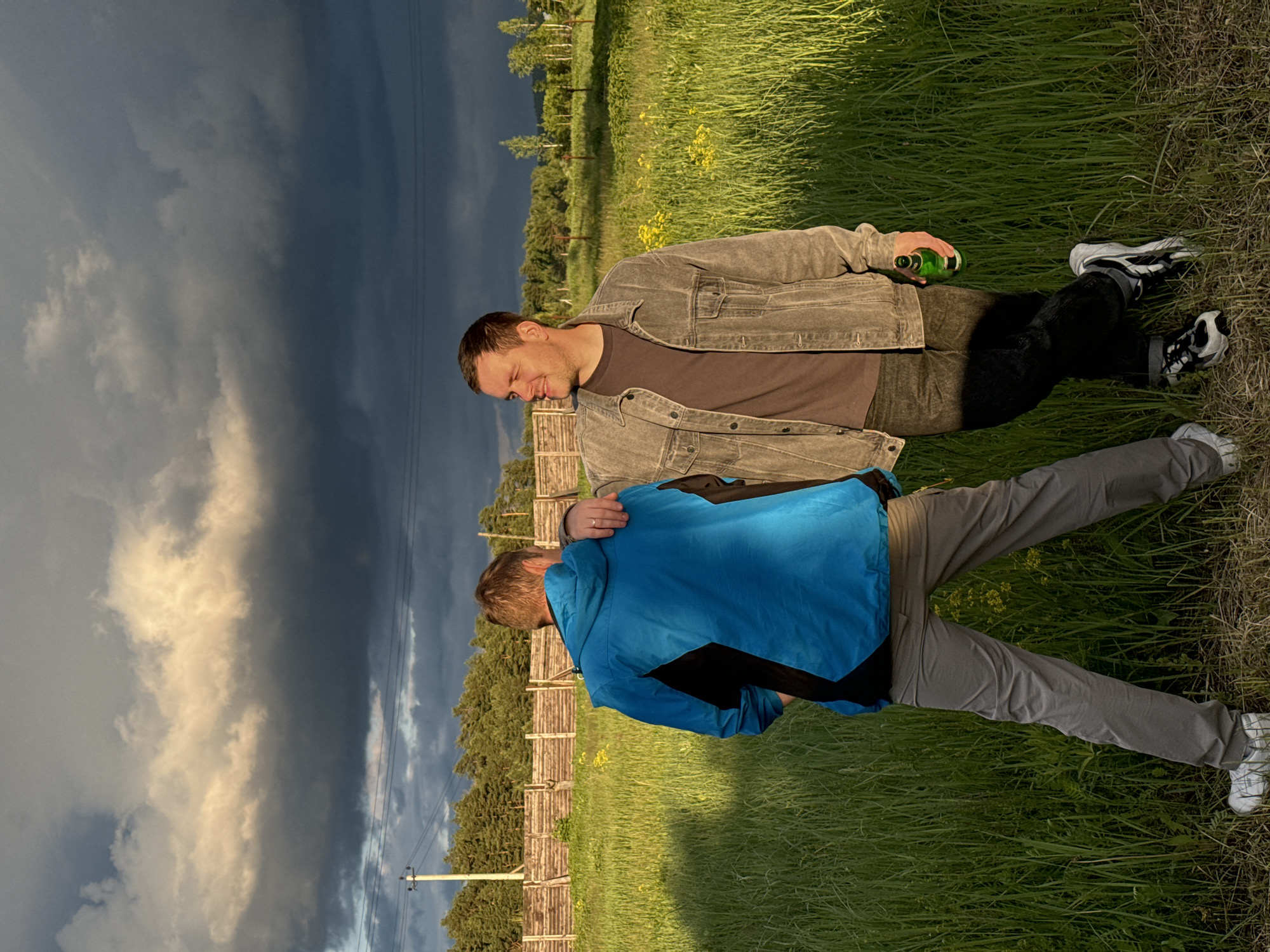 Two men are lying on a grassy field in Vysokinichi, Russia. One man wears a grey jacket, brown shirt, and black pants, holding a green bottle. The other man wears a blue and black jacket, and grey pants. The man in the grey jacket has his hand on the other man's shoulder. A wooden fence runs along the edge of the field. A dark, cloudy sky fills the background. Power lines and a utility pole are visible.