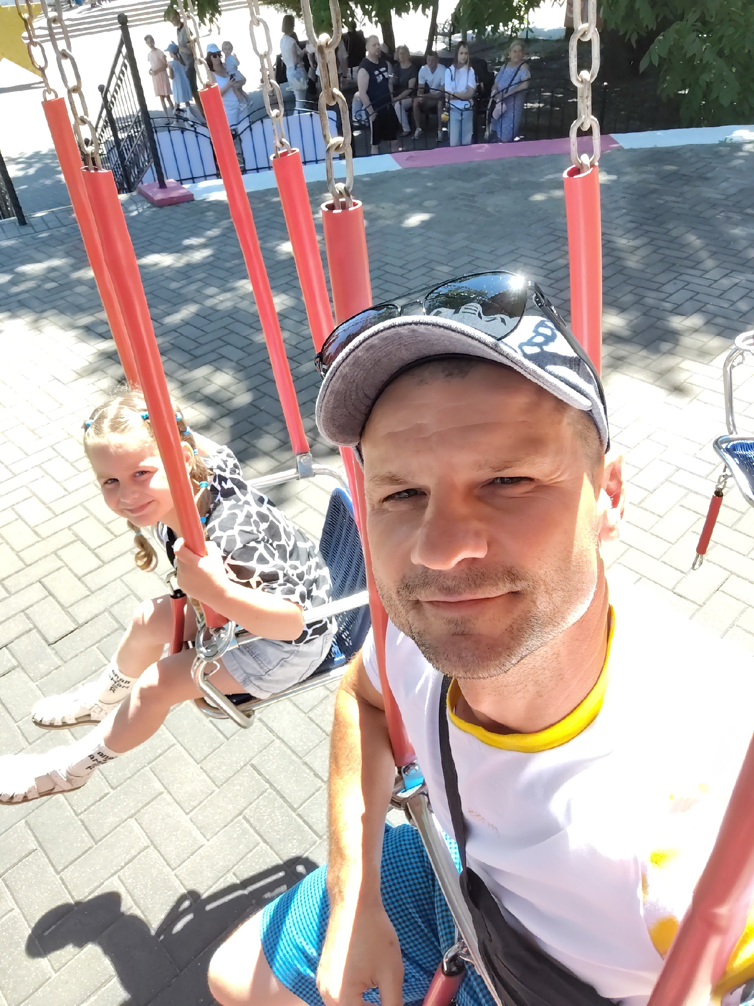 In Homyel, Belarus, on a sunny day, a man and a young girl are on a swing set. The man, wearing a cap and sunglasses, is taking a selfie. The girl, with her hair in braids, is smiling and looking at the camera. They are on a paved area with trees in the background. Other people are visible in the distance, some sitting on benches. The swing set has red metal poles and chains.