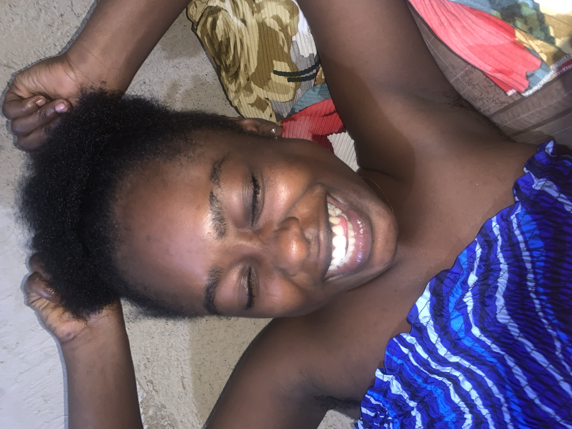 A young Black woman with her eyes closed and mouth open in a wide smile is lying on her back. Her hands are raised above her head, fingers intertwined, and resting on her dark, curly hair. She is wearing a blue and white striped top. The background consists of a textured wall and patterned fabric. The scene appears to be indoors in Rafin Par, Nigeria.