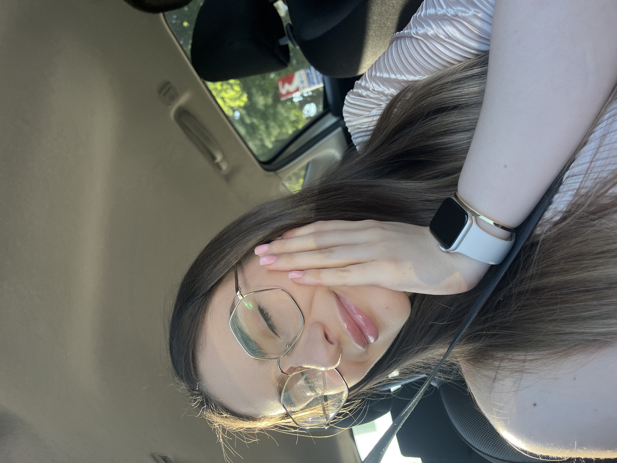 A young woman is sitting in a car. She is wearing glasses and a smart watch. Her hand is covering part of her face.