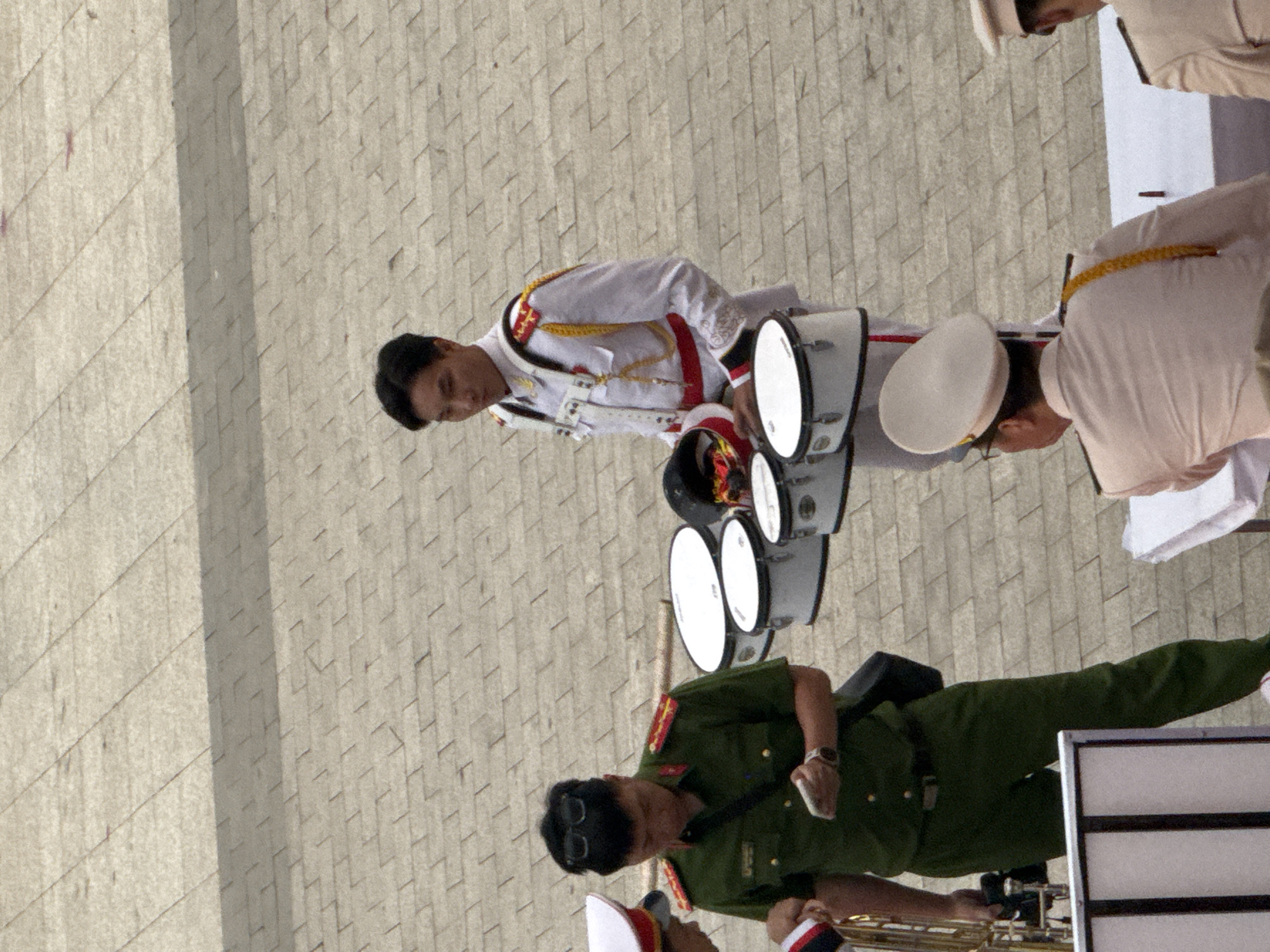 The image depicts a street scene in Da Lat, Vietnam, likely during a formal event or parade. In the foreground, a person in a green military uniform with sunglasses is holding a smartphone and a watch is visible on their wrist. They are standing near a brass instrument and several drums. To their right, a person in a white formal uniform with red and gold accents is holding a drumstick and a drum. Behind them, two individuals in white uniforms with hats are partially visible. The ground is paved with grey interlocking bricks.