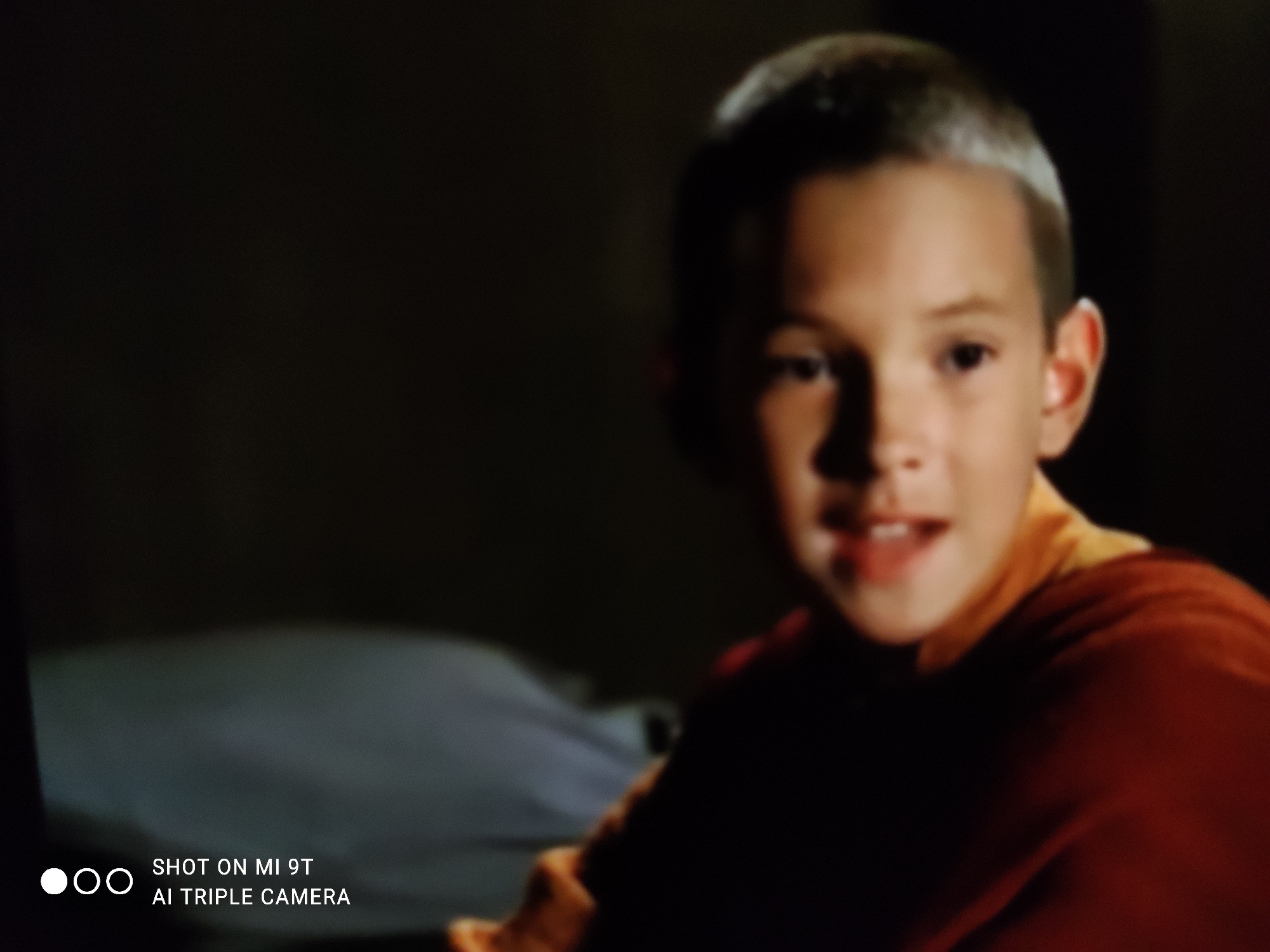 The image is a close-up, slightly blurry, indoor shot of a young boy with short, light brown hair. He is wearing an orange and yellow garment, possibly a robe or shirt. His facial expression appears to be one of mild surprise or inquiry, with his mouth slightly open. The background is dark and indistinct, suggesting a dimly lit room. The bottom left corner of the image contains a watermark that reads "SHOT ON MI 9T AI TRIPLE CAMERA". There is no discernible information within the image that suggests a location in Khazar Raion, Azerbaijan.